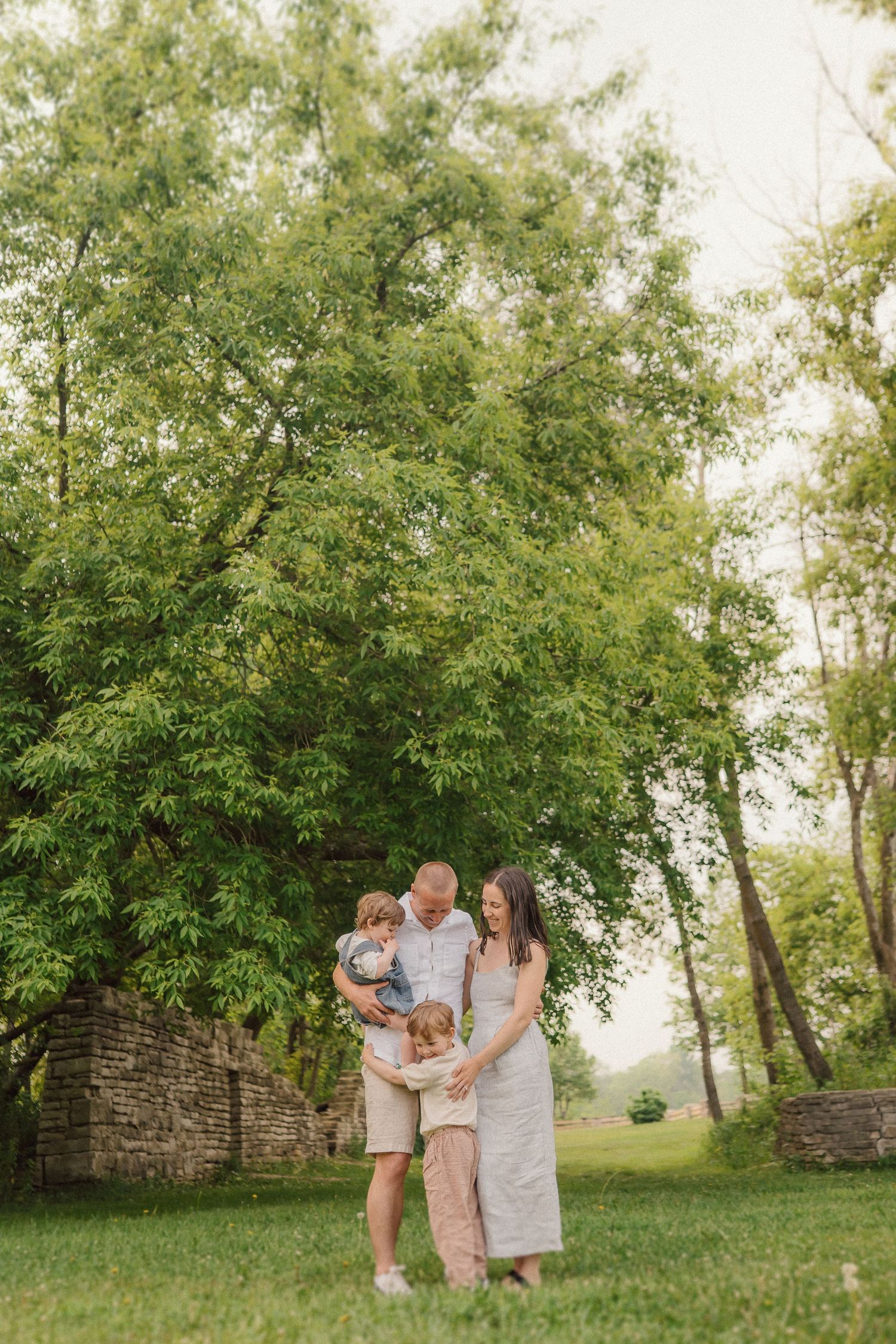 A family shares a tender moment together while standing under tall trees near a stone wall.