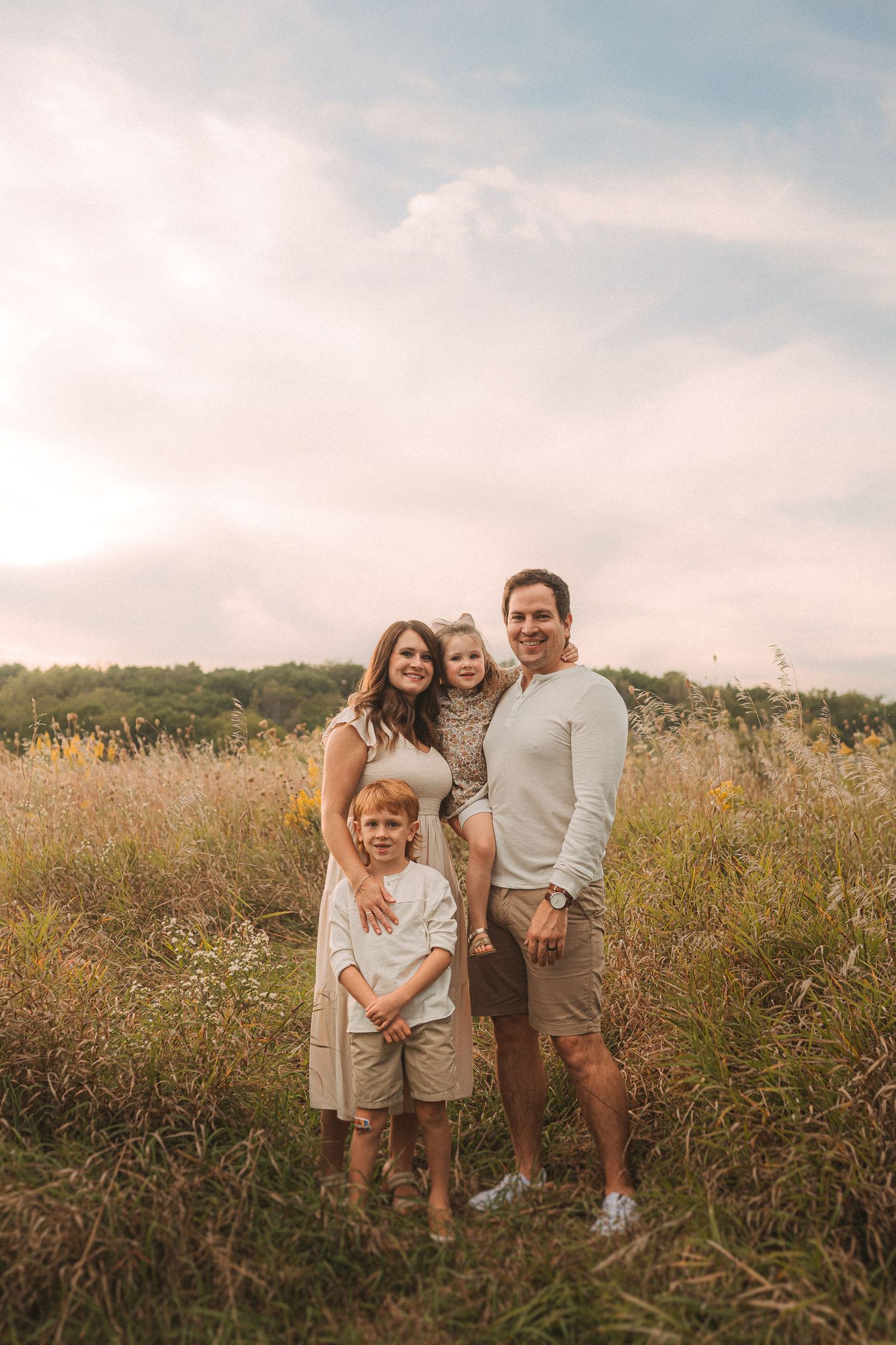 A family stands together in a sunlit field of tall grass during golden hour at sunset.