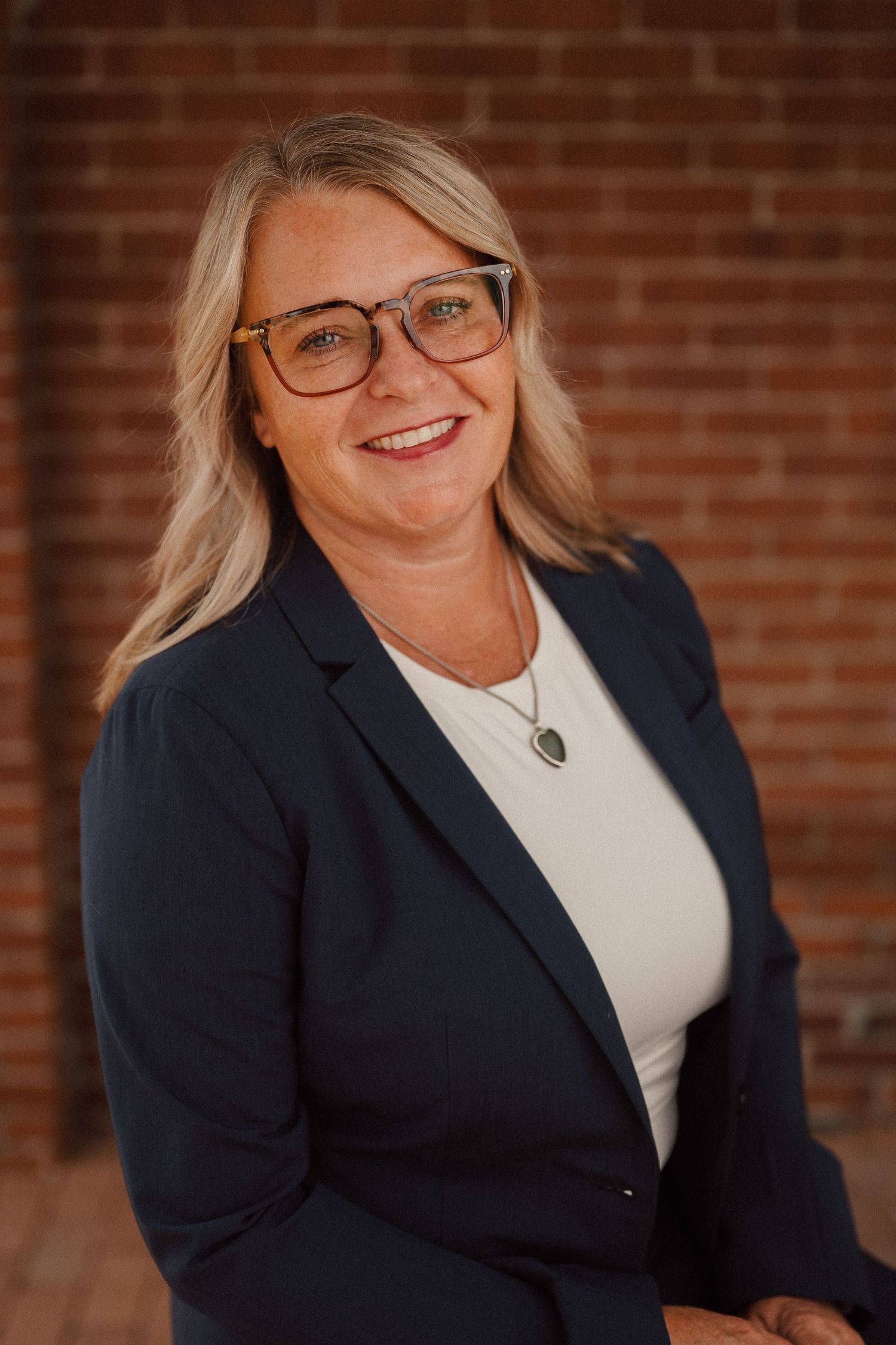 Professional headshot against brick wall showing person in navy blazer and white top smiling warmly.