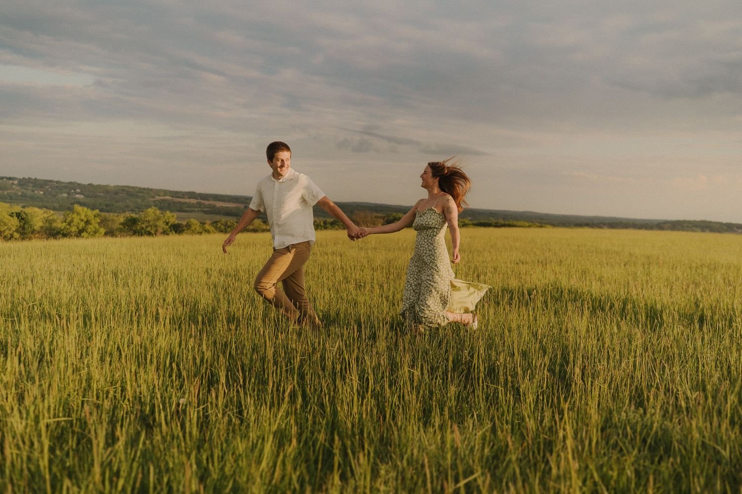 A couple dances playfully in a sunlit meadow with tall grass and dramatic clouds overhead.