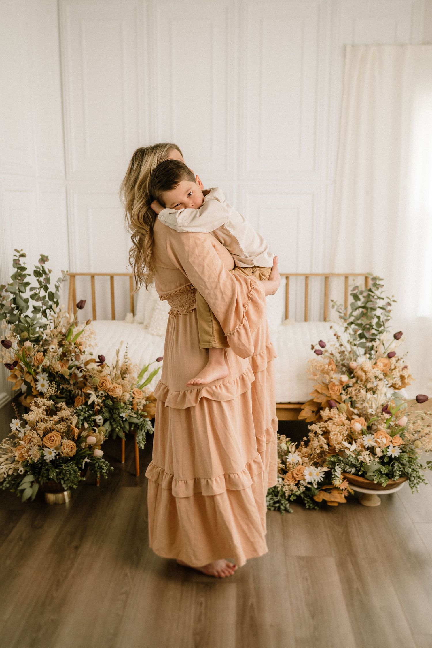 A tender moment in a peachy-pink ruffled dress against a backdrop of dried floral arrangements and wooden furniture.