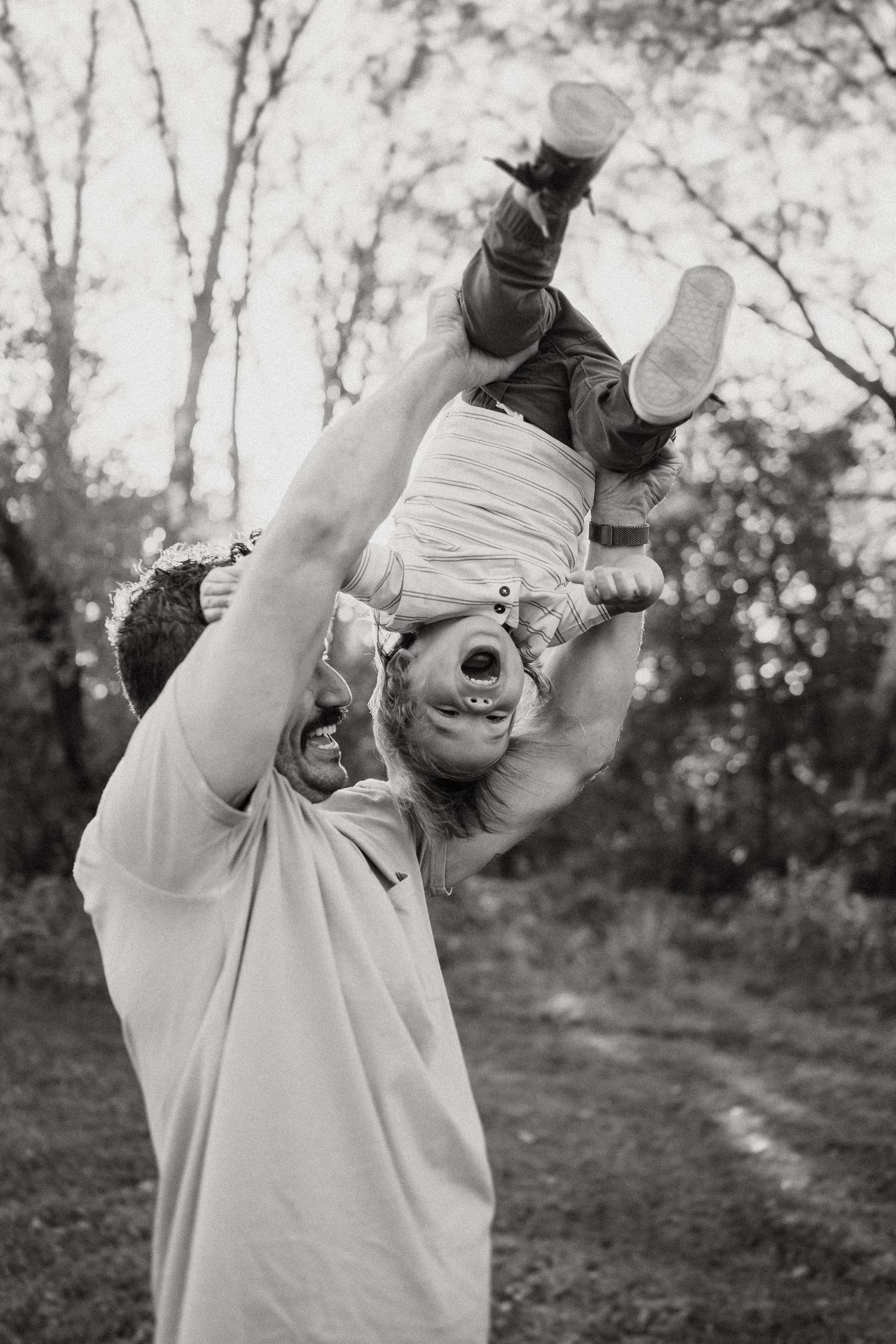 Black and white capture of joyful playtime as parent lifts child upside down outdoors.