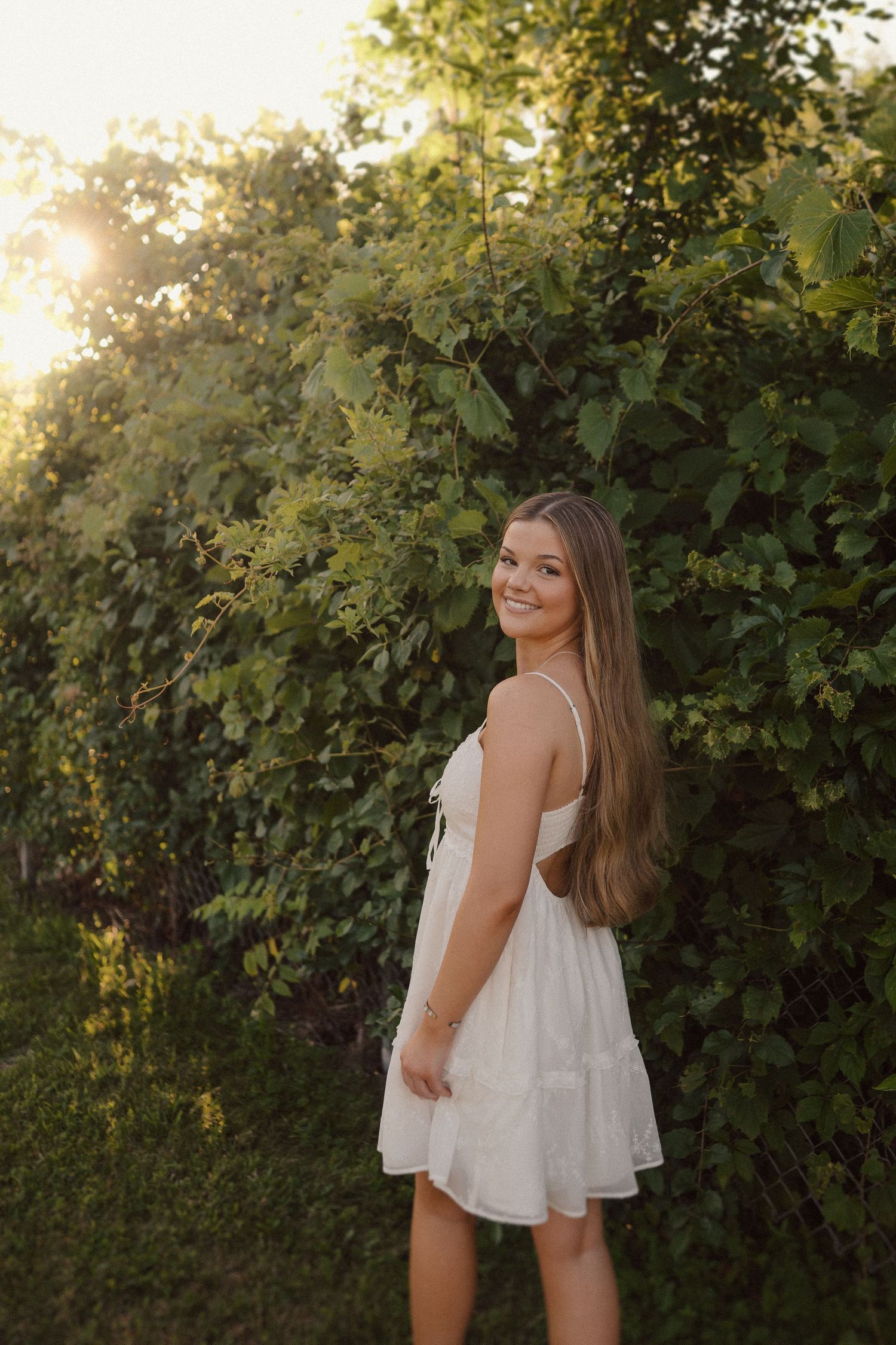 Person in white sundress poses against lush greenery during golden hour lighting.