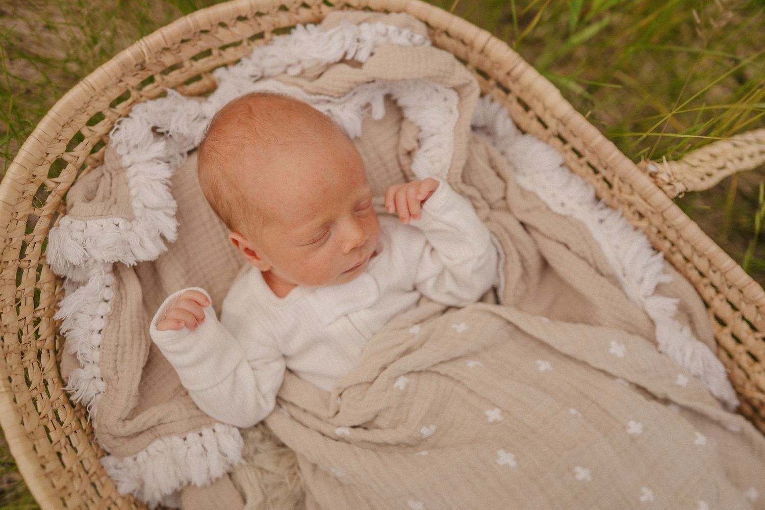 Sleeping newborn in white outfit lying peacefully in woven basket outdoors.