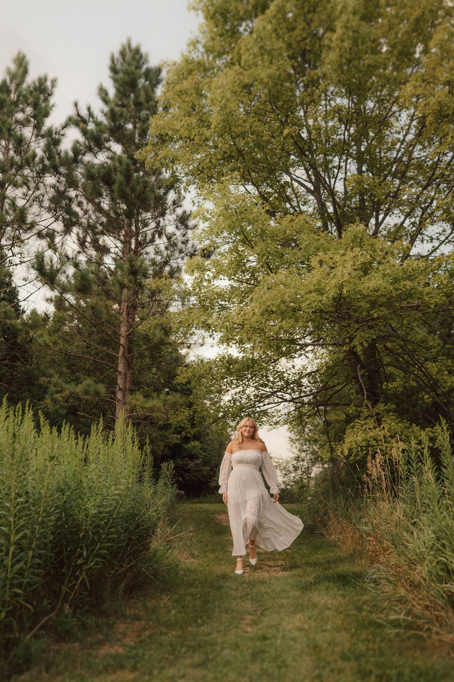A graceful figure in a flowing white dress walks through a verdant garden path lined with trees and greenery.