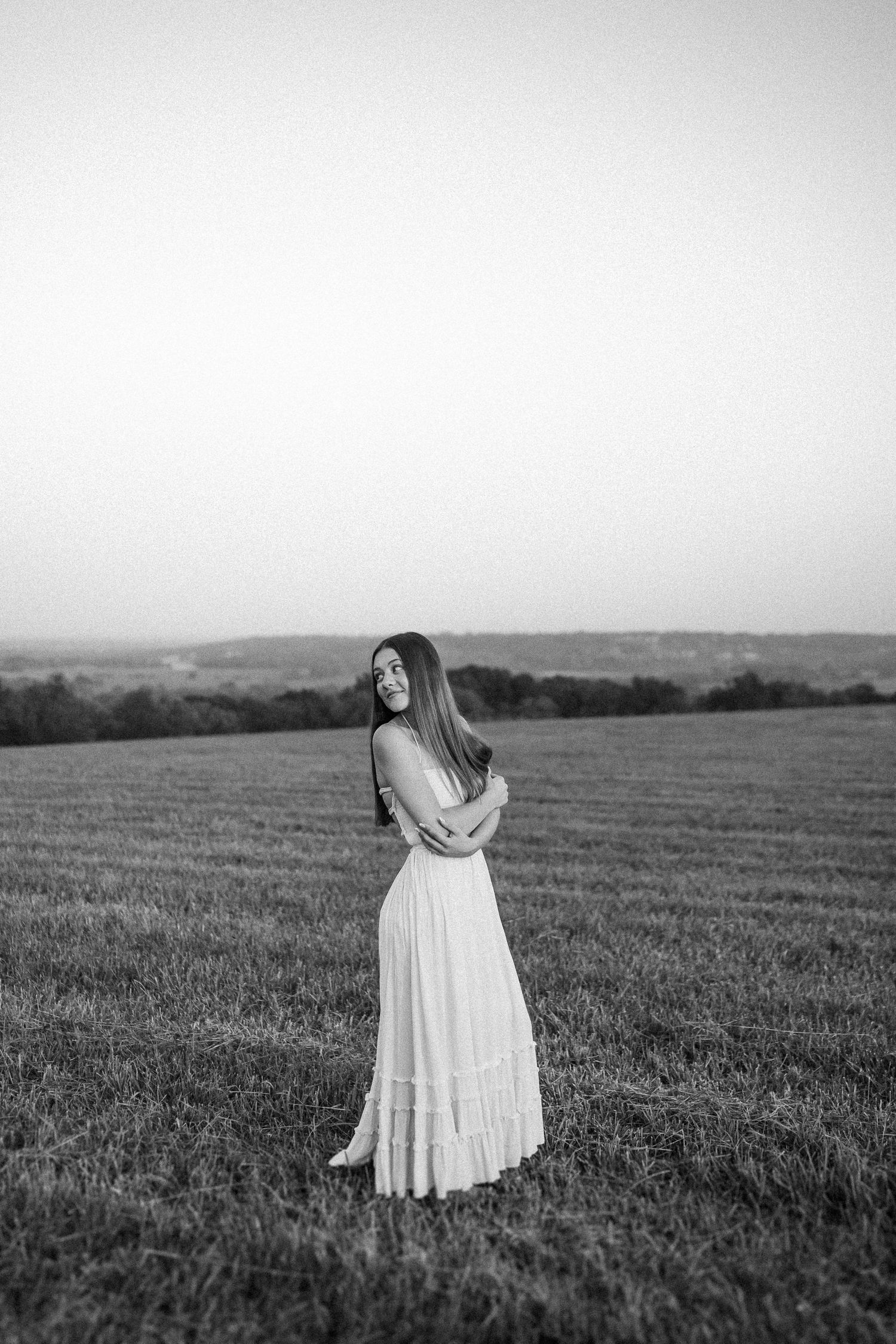 A black and white photo shows a figure in a white dress standing in a vast open field at dusk.
