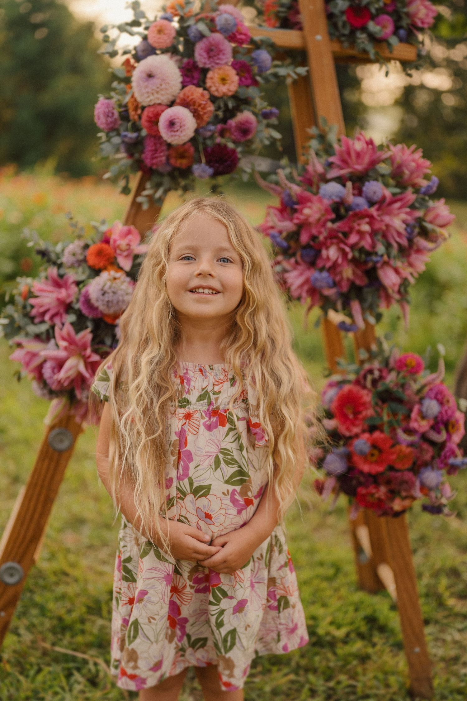 A young child in a floral dress smiles in front of a wooden ladder decorated with pink and purple flower arrangements.