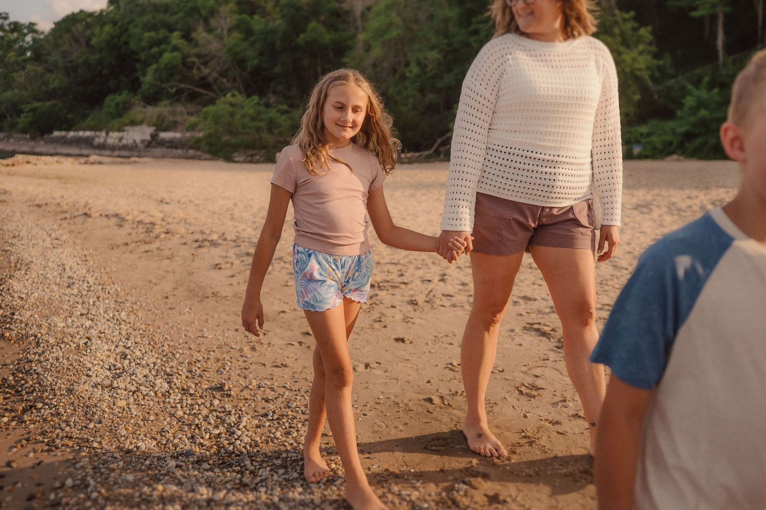 People walking along a rocky beach shore with trees in the background at sunset.