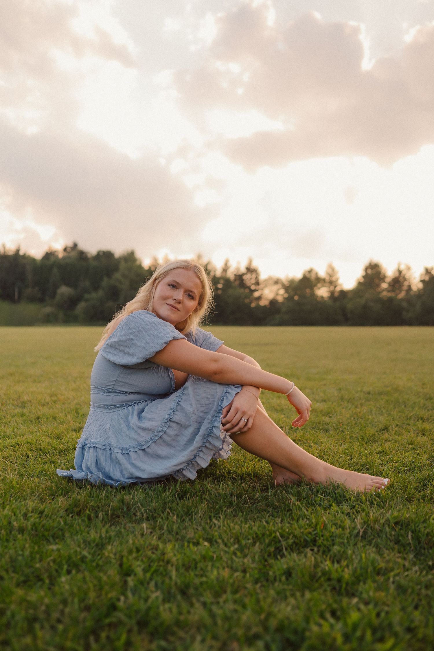 A woman in a blue dress relaxes on a grassy field during golden hour sunset.