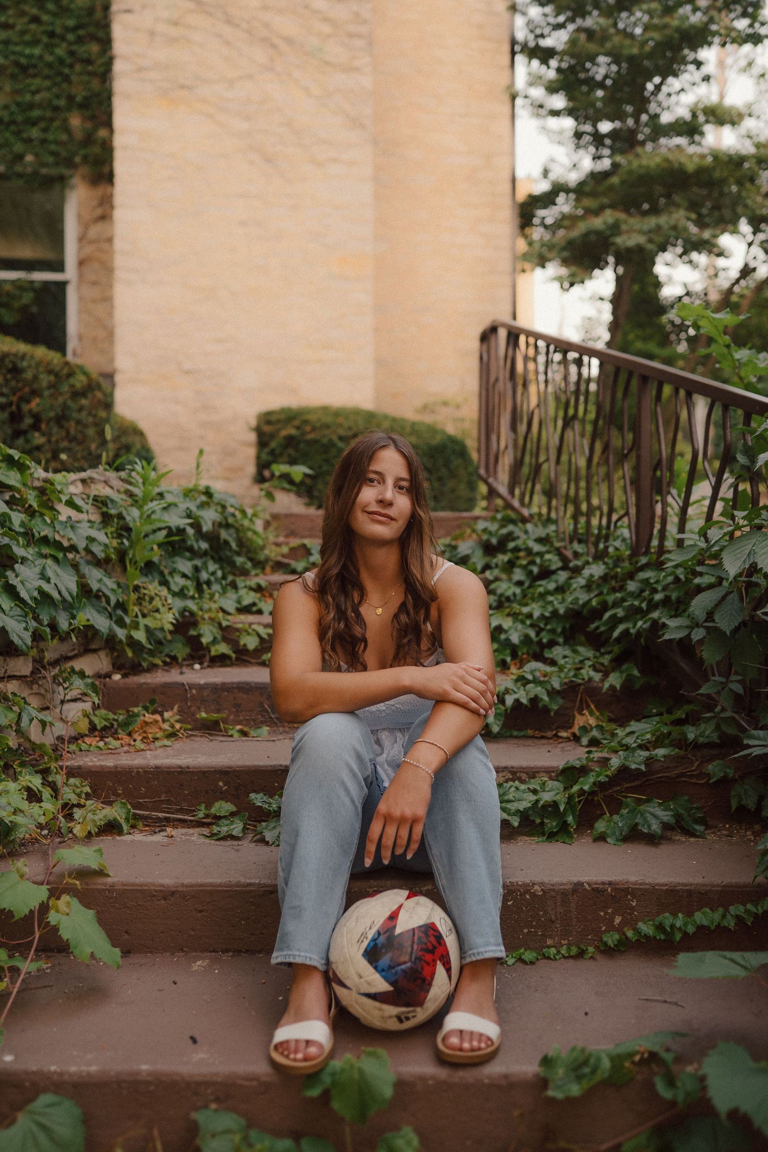A person in jeans sits on outdoor steps with soccer ball and ivy covered walls.