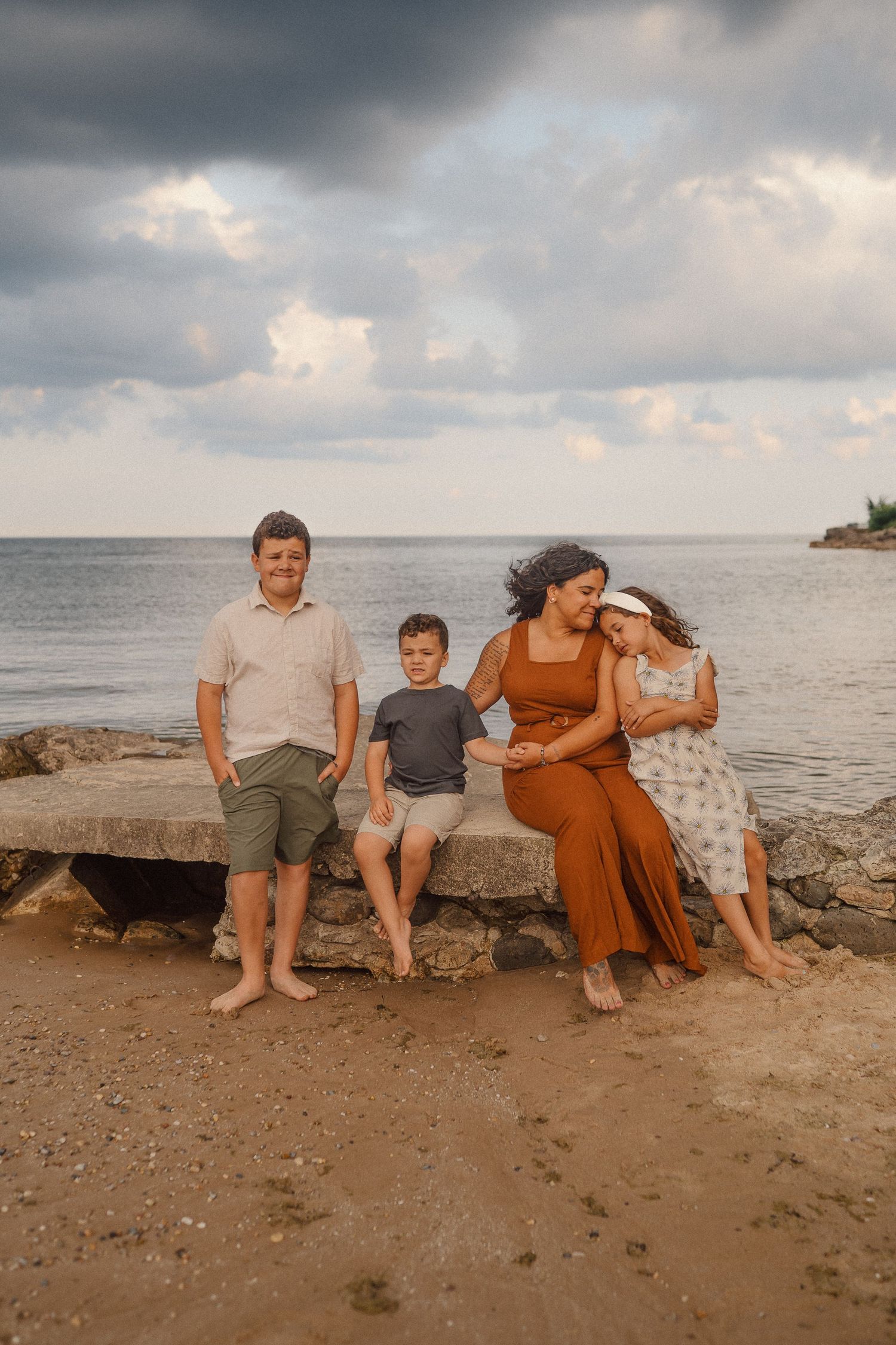 A family sits together on rocky beach shoreline at sunset with moody clouds overhead.