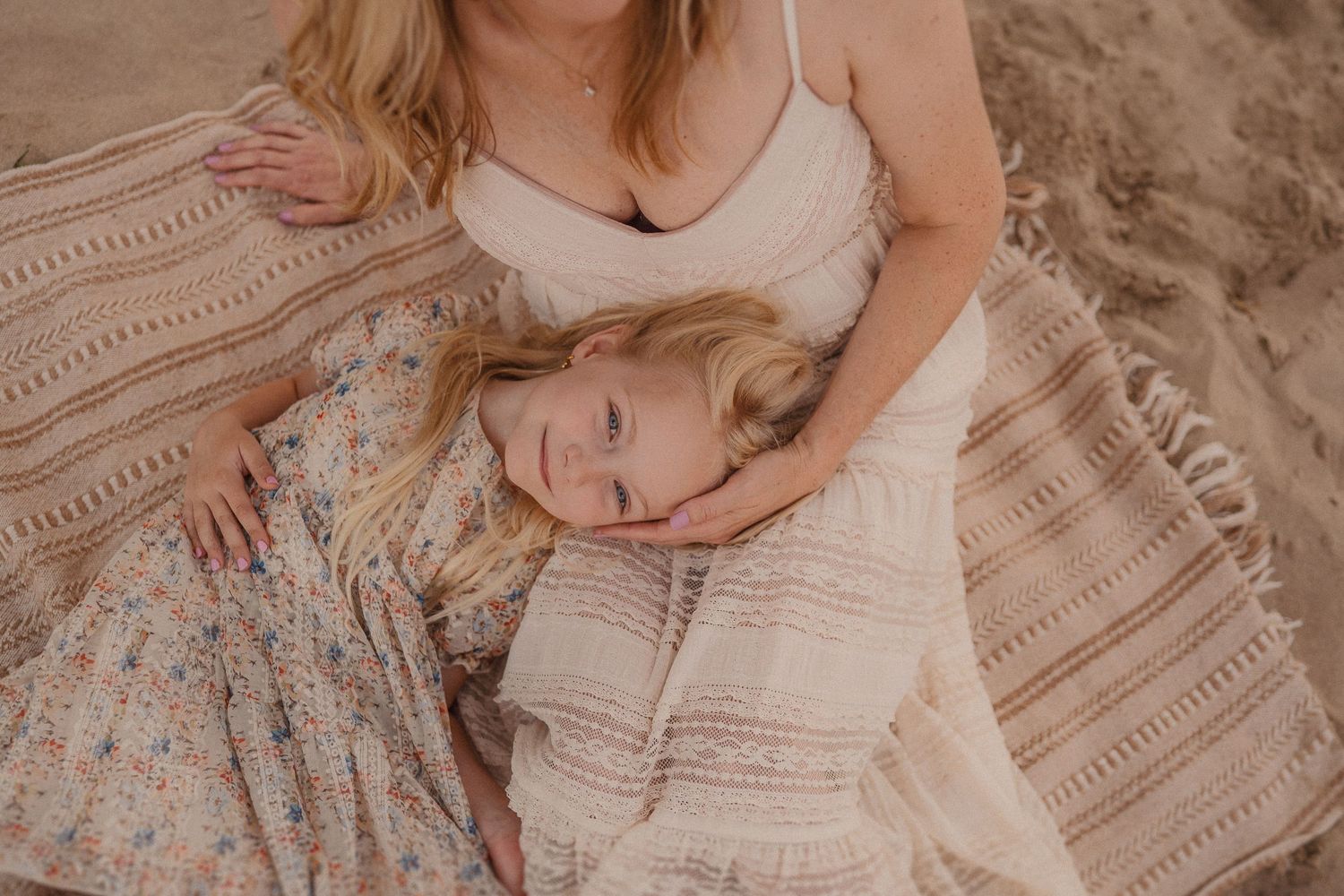 A tender moment captured on a striped blanket shows two people in light flowy dresses resting peacefully on sand.