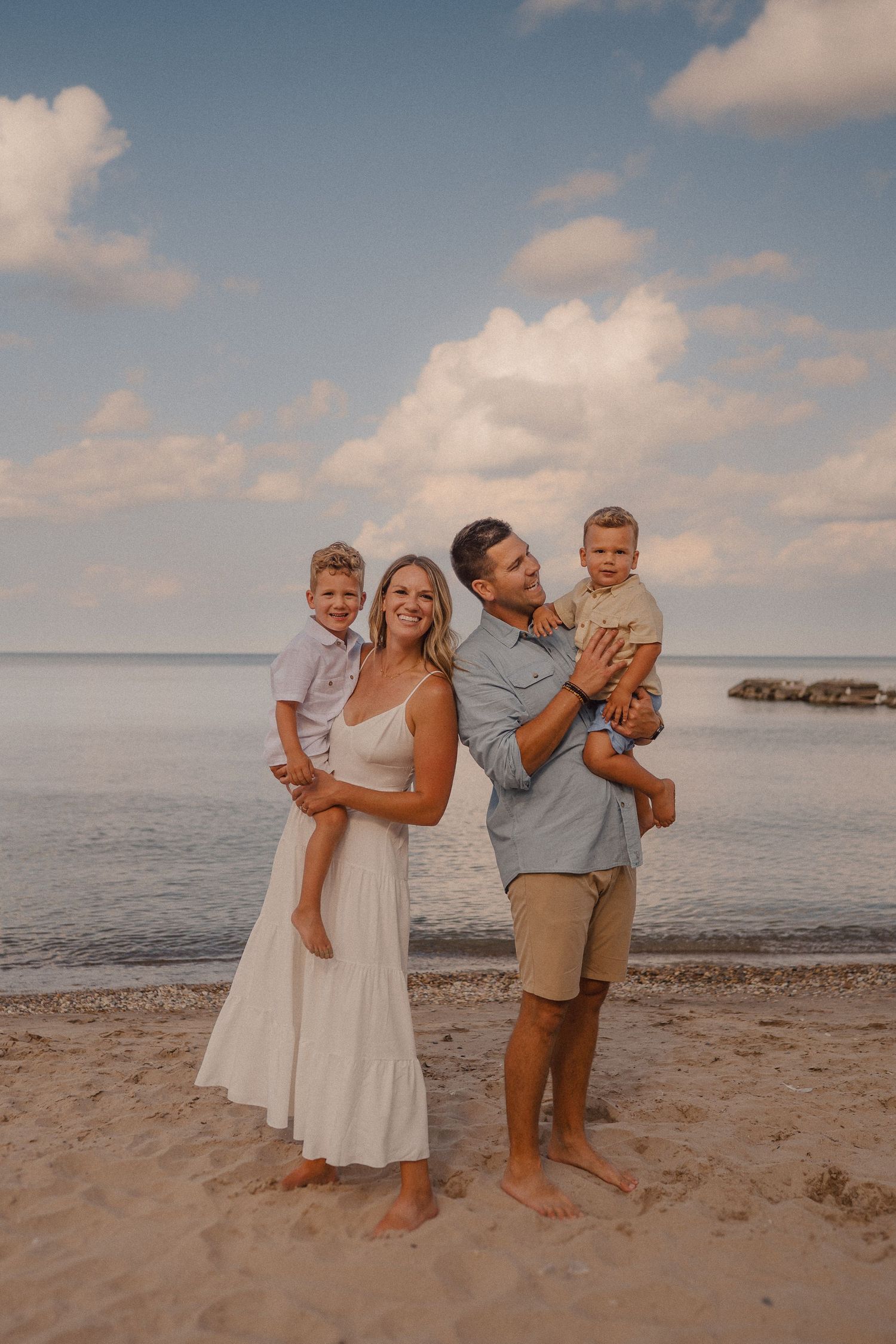 A family stands together on a sandy beach at sunset with the ocean and cloudy sky in the background.