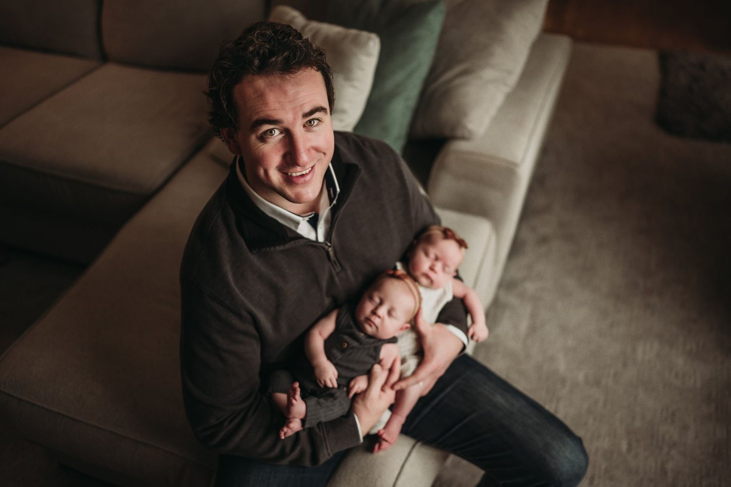 Parent on couch smiles joyfully while holding content newborn baby in casual home setting.