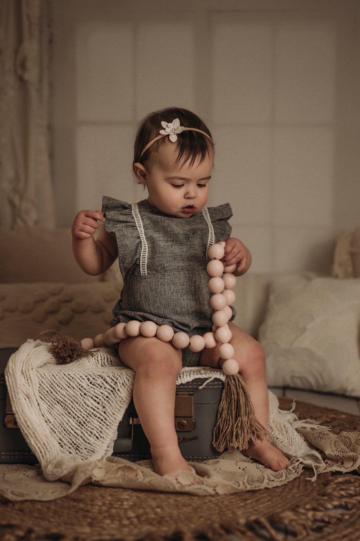 A small child in gray romper playing with wooden beads while sitting on vintage suitcase.