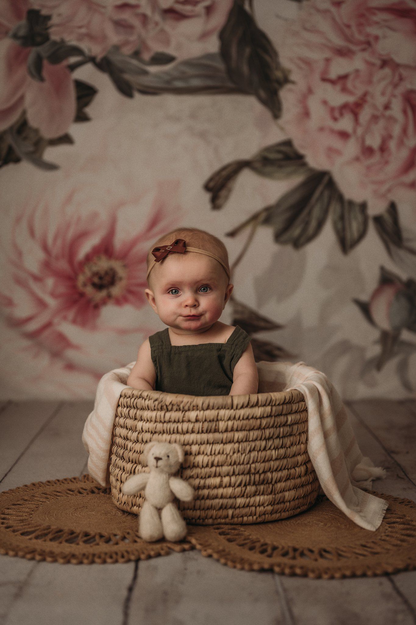Child in olive romper sits in woven basket with decorative floral background.