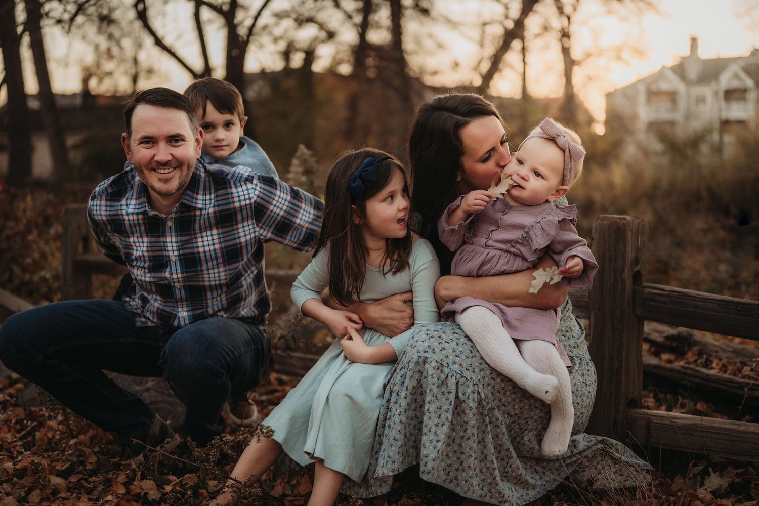 A loving family shares laughter while sitting on a wooden fence at sunset.