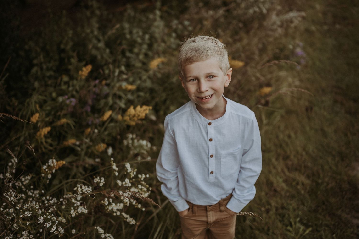 Gentle outdoor portrait of a child wearing white shirt and brown pants against a nature backdrop.