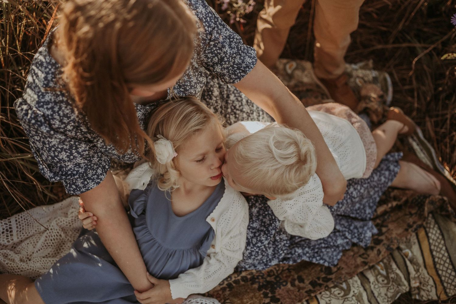 Tender family moment captured on blanket in field during golden hour.