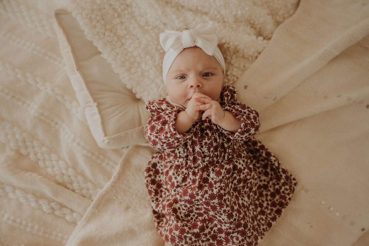 Baby in floral dress and white bow lying on cream colored blankets.