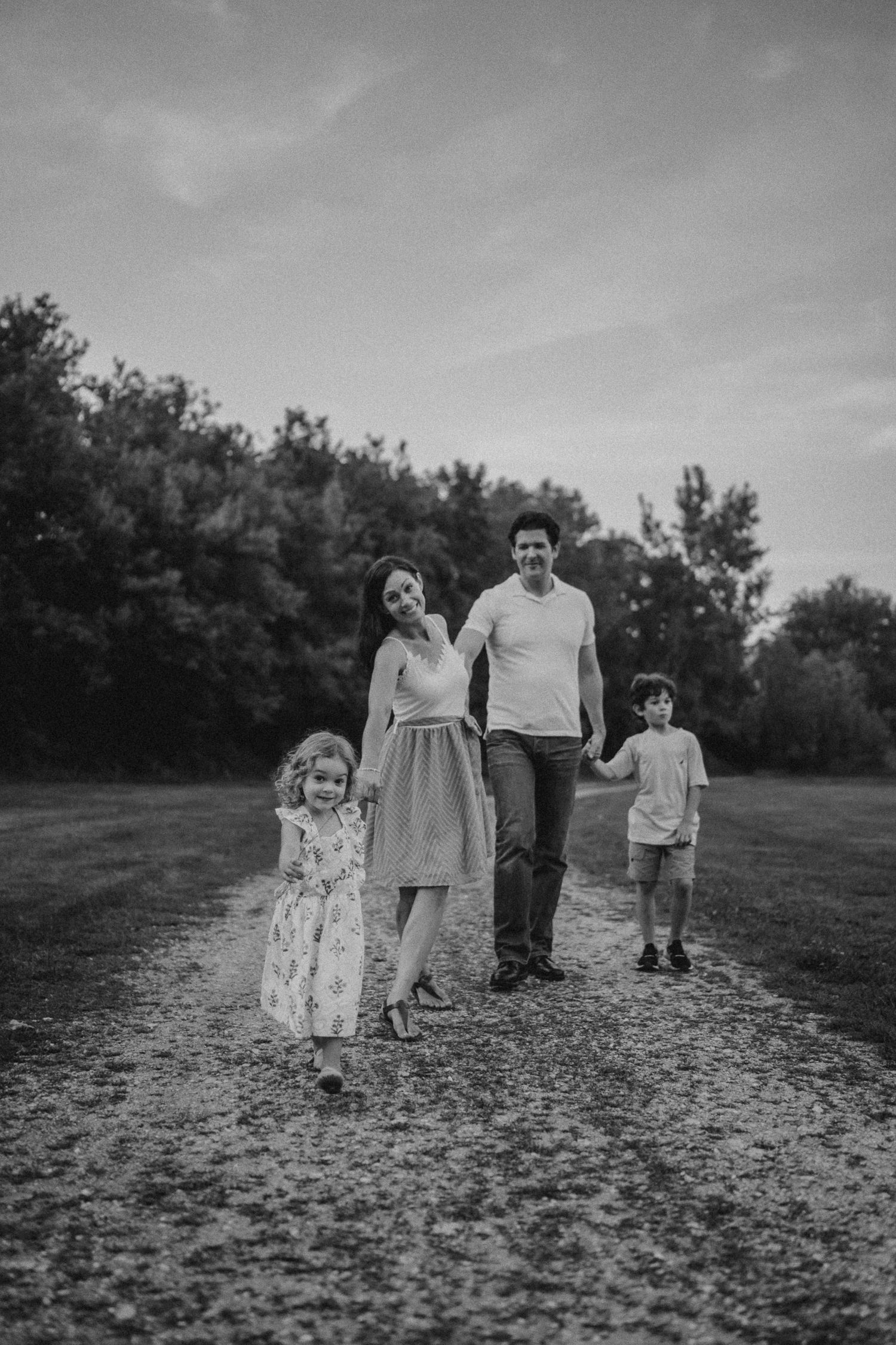 A family walks together on a dirt path through a grassy field at dusk in black and white.
