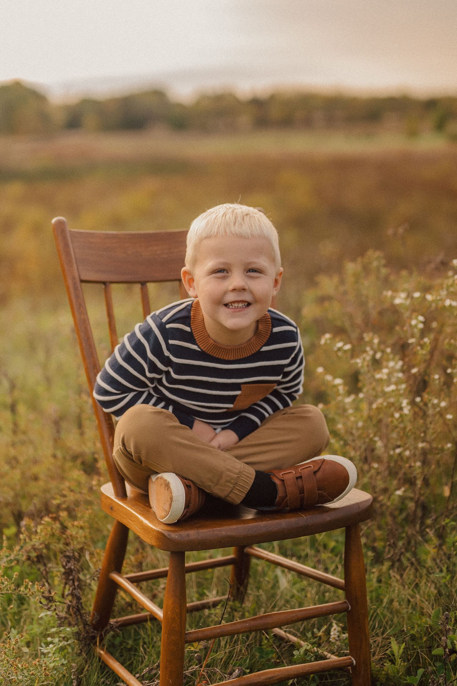 Child sitting cross-legged on wooden chair in field with wildflowers during sunset.
