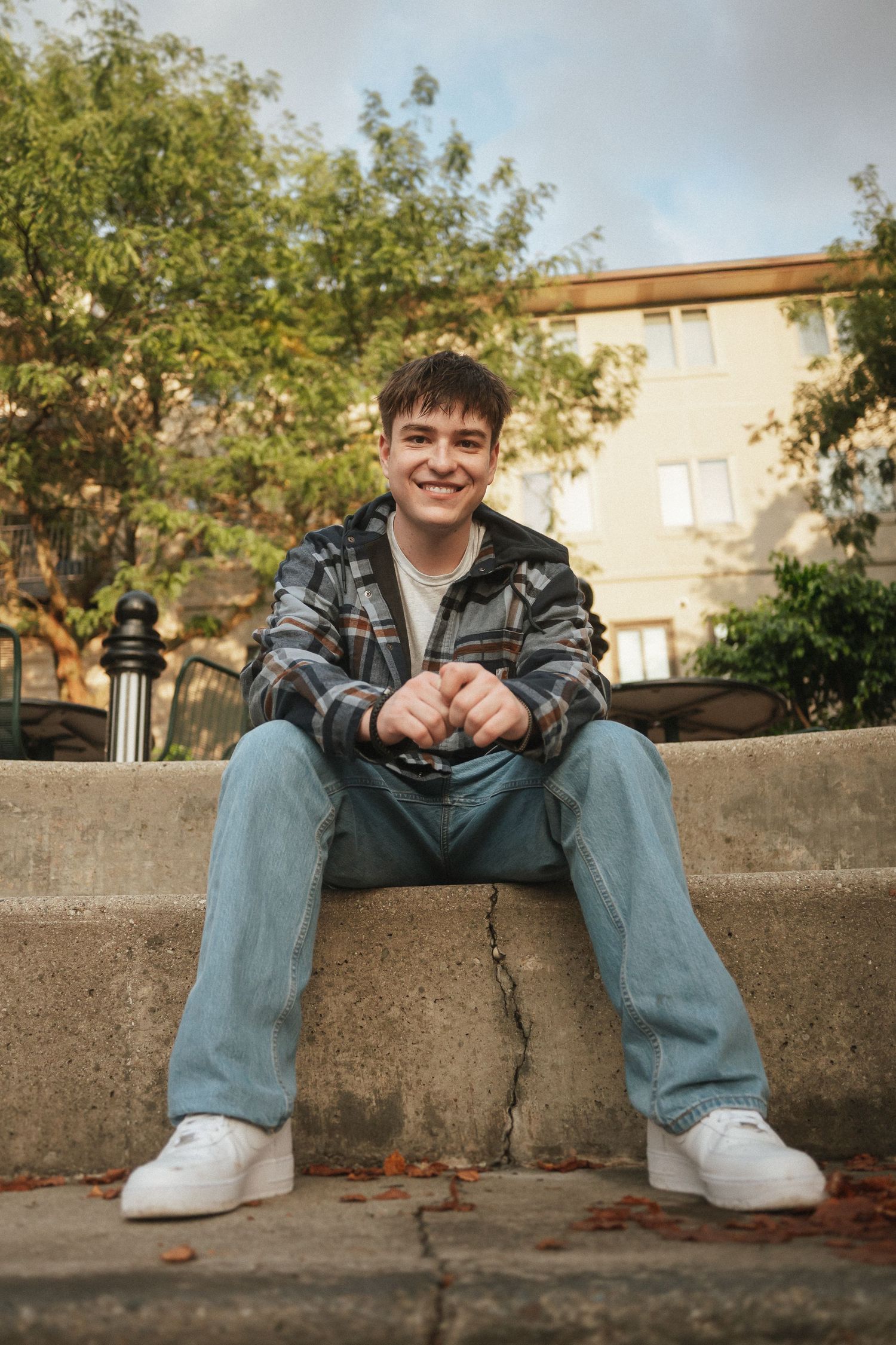 Person in a camo jacket and jeans sits casually on concrete steps with trees and building in background.