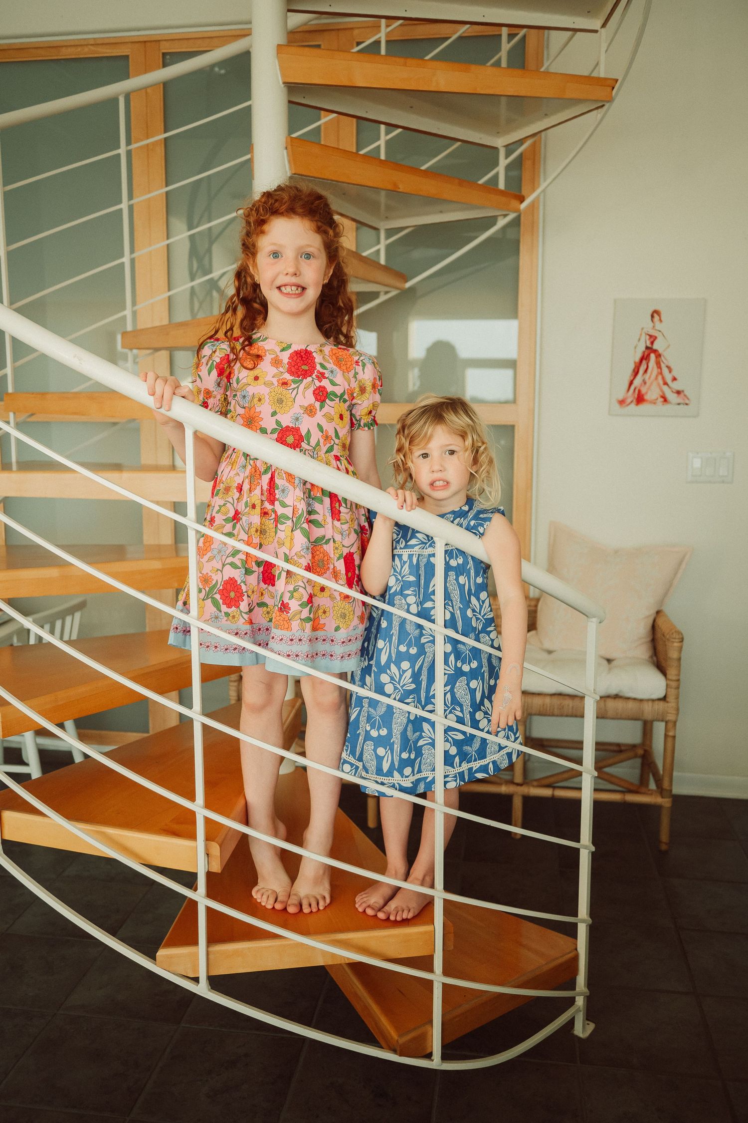 Two children in colorful summer dresses posing on white stairs indoors.