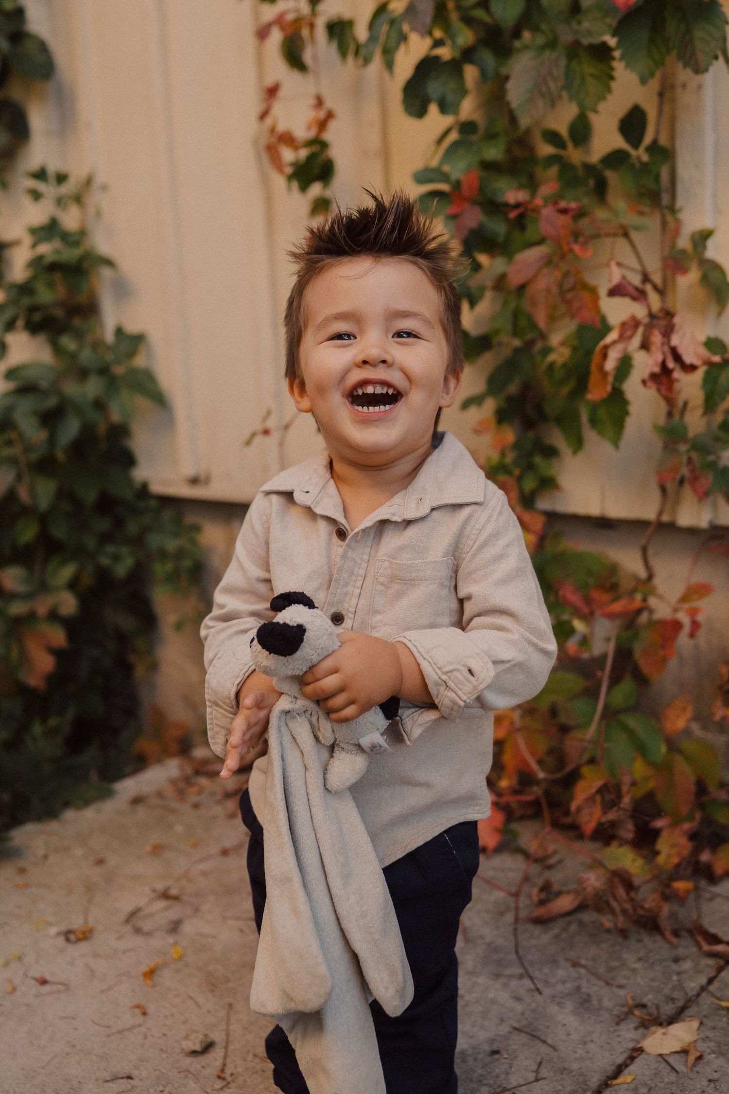 A child laughs while holding a plush toy bunny outdoors against an autumn backdrop with red ivy leaves.