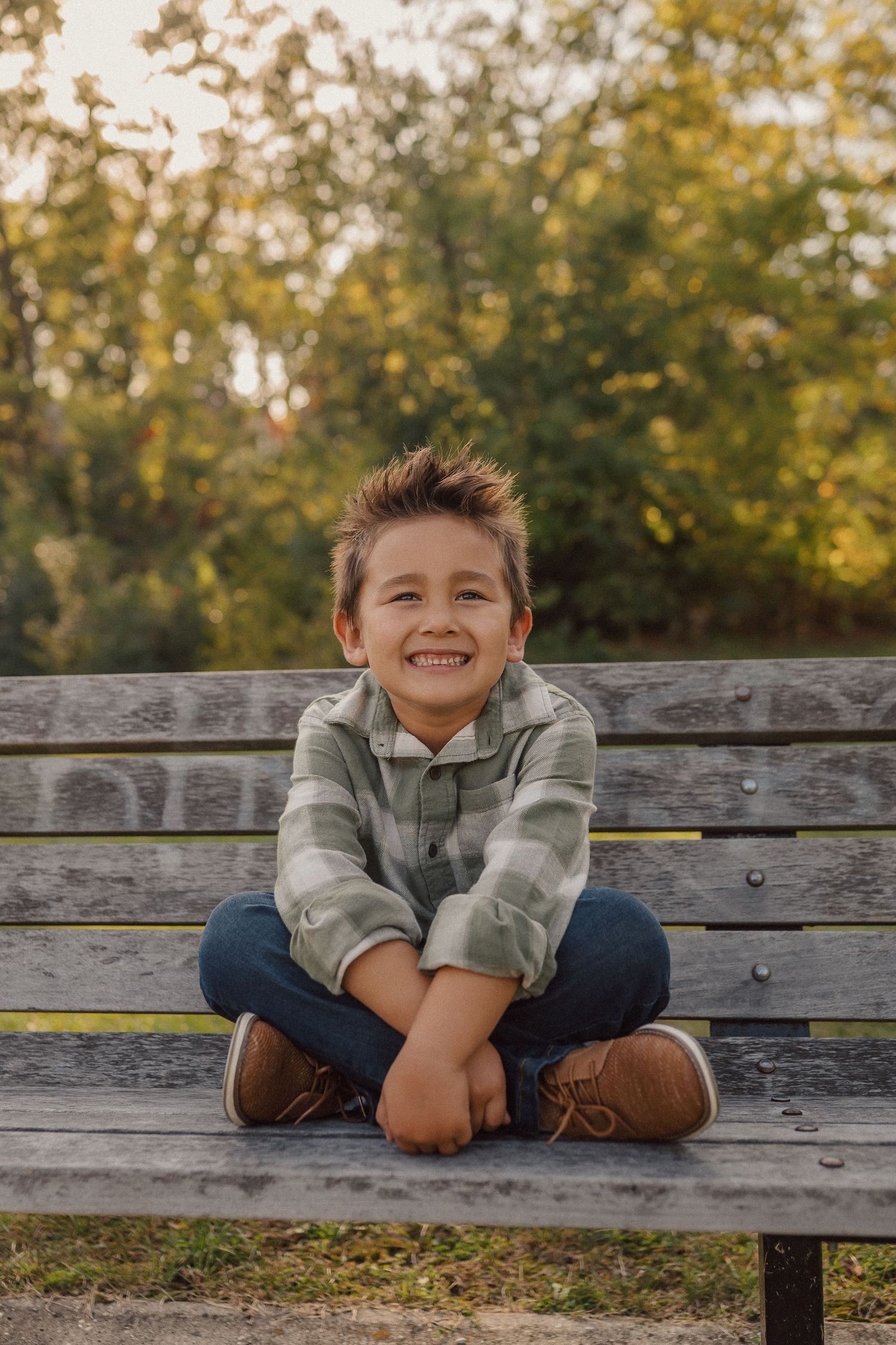 A young child sits casually on a wooden bench wearing jeans and a light jacket on an autumn day.
