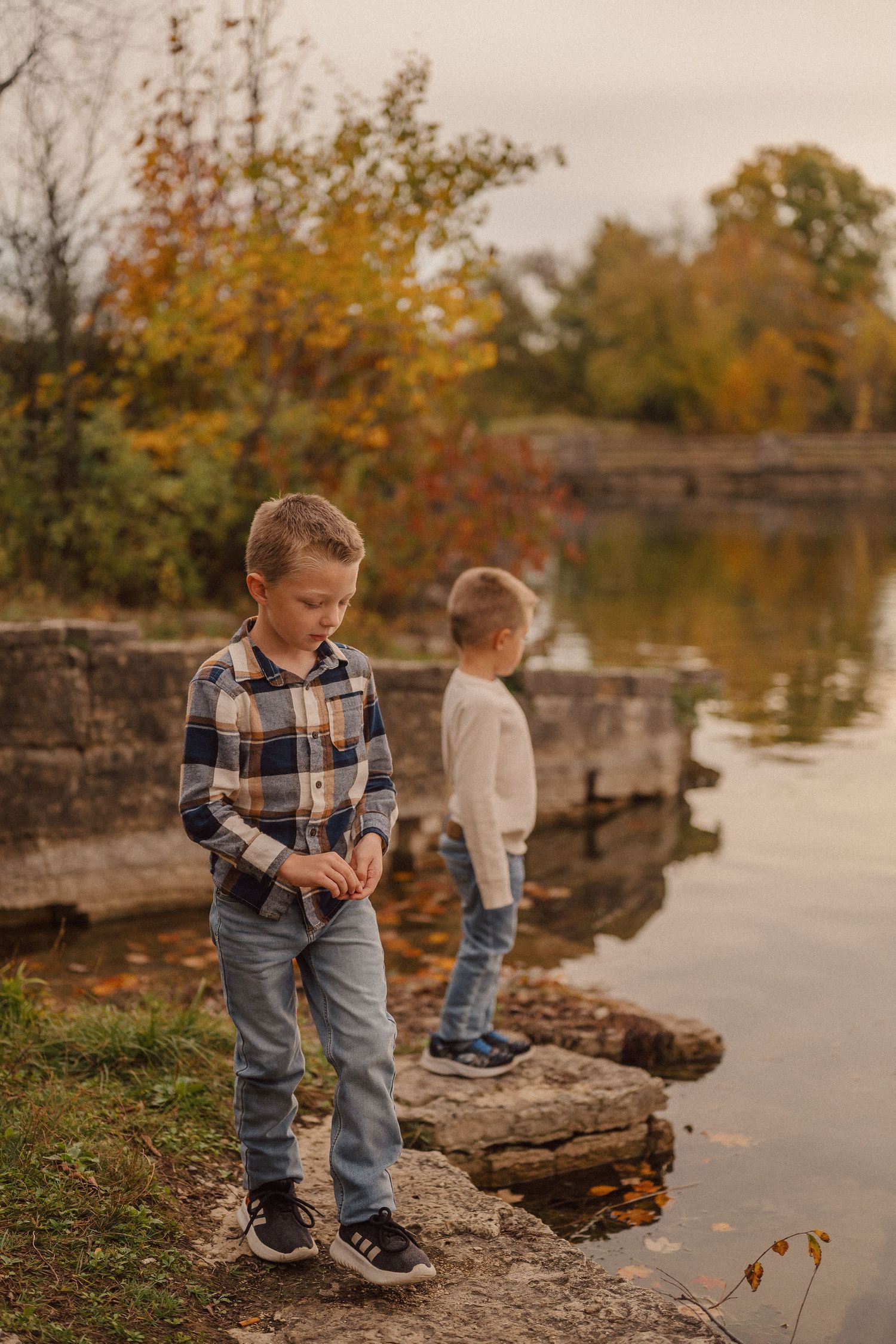 Children exploring rocky ledges by a calm lake during fall with golden leaves in background.