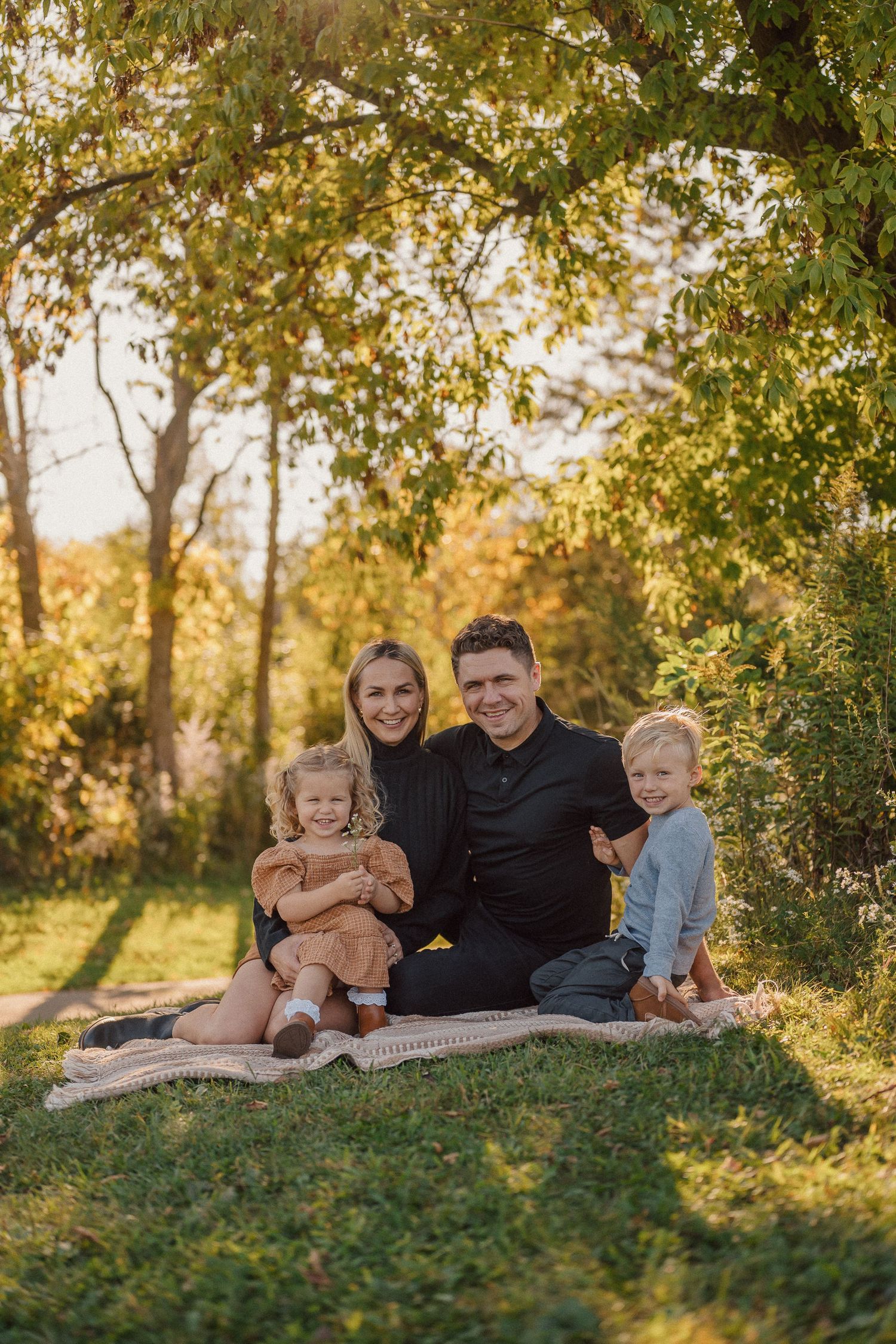 Family relaxing together on grassy hill during golden hour with autumn trees creating a natural backdrop.