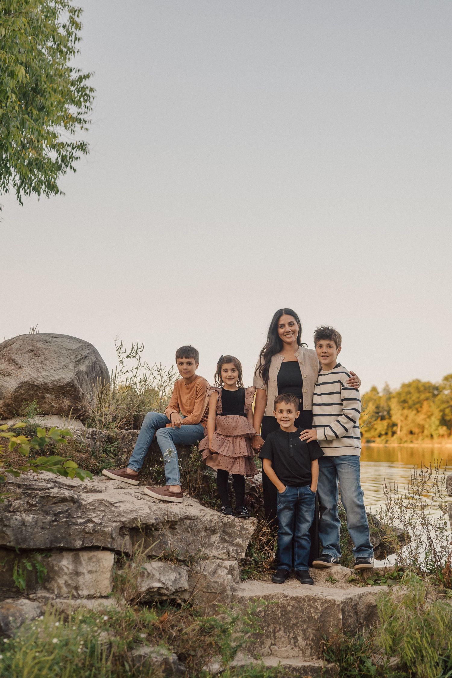Family group sitting together on large rocks by a lake with autumn trees in the background during sunset.