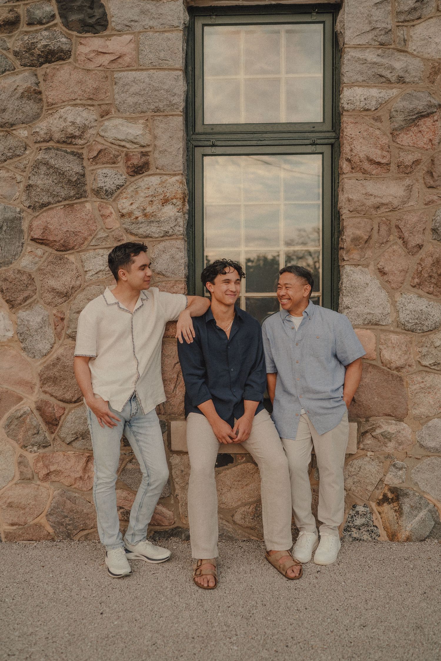 Three young men pose and laugh together outside a rustic stone building in warm evening light.
