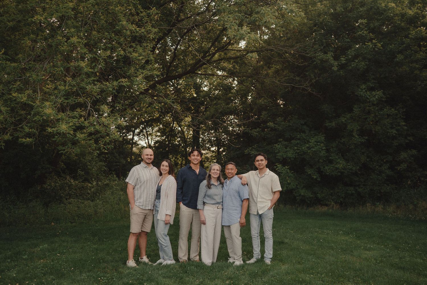 Family of six posing together on green grass lawn surrounded by lush trees during golden hour.