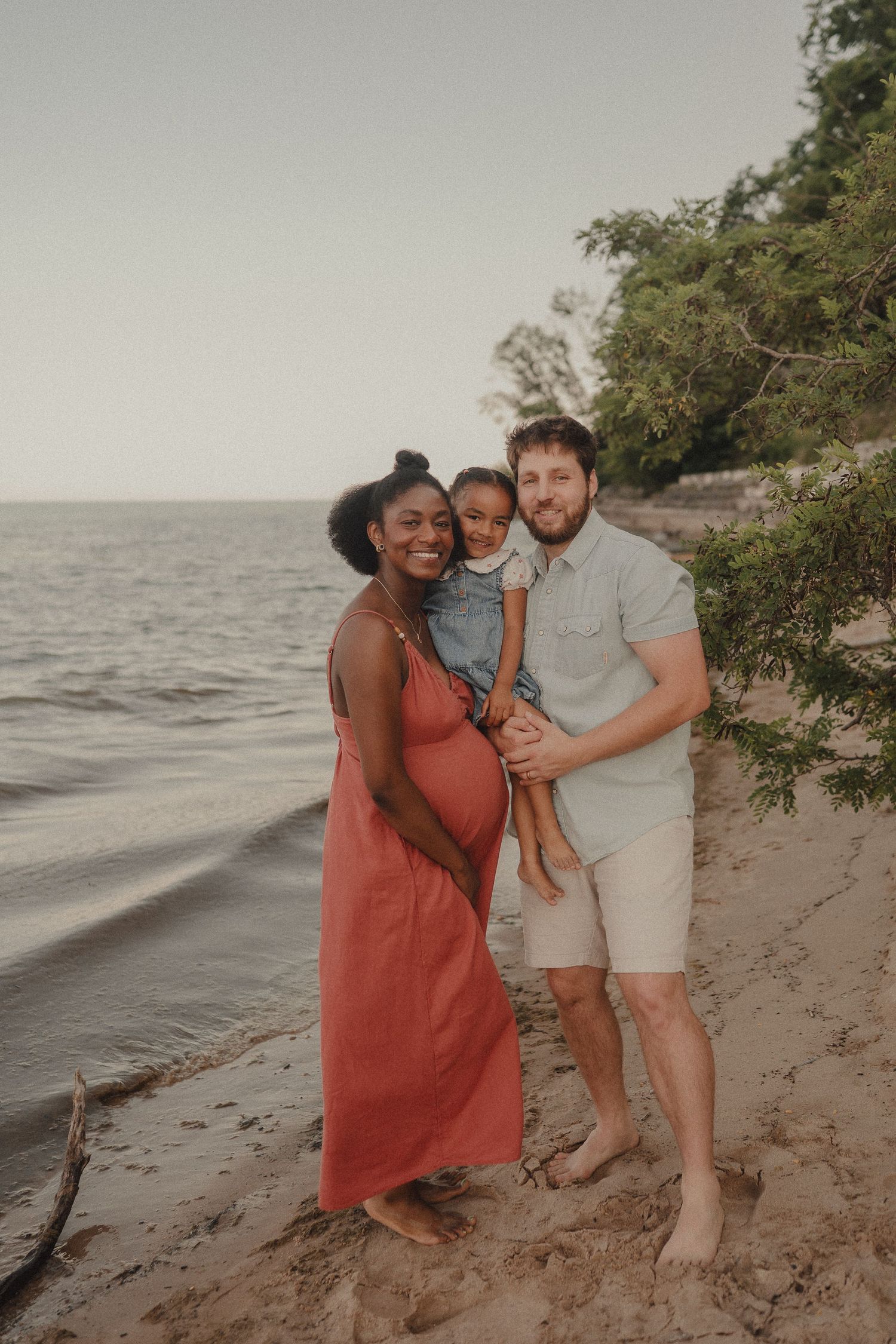 Interracial family maternity beach portrait with pregnant mother in coral dress, father holding toddler by lakeside.