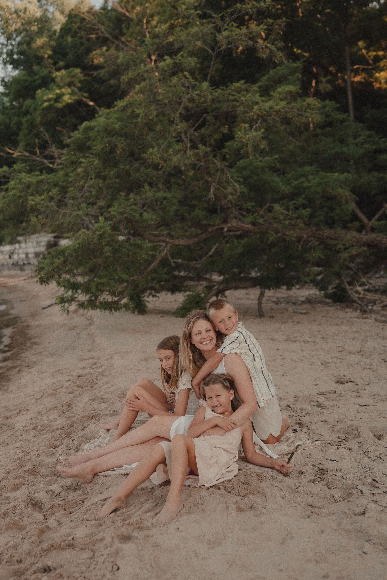 A smiling mother sits on sandy beach with three children hugging her near lush green trees.