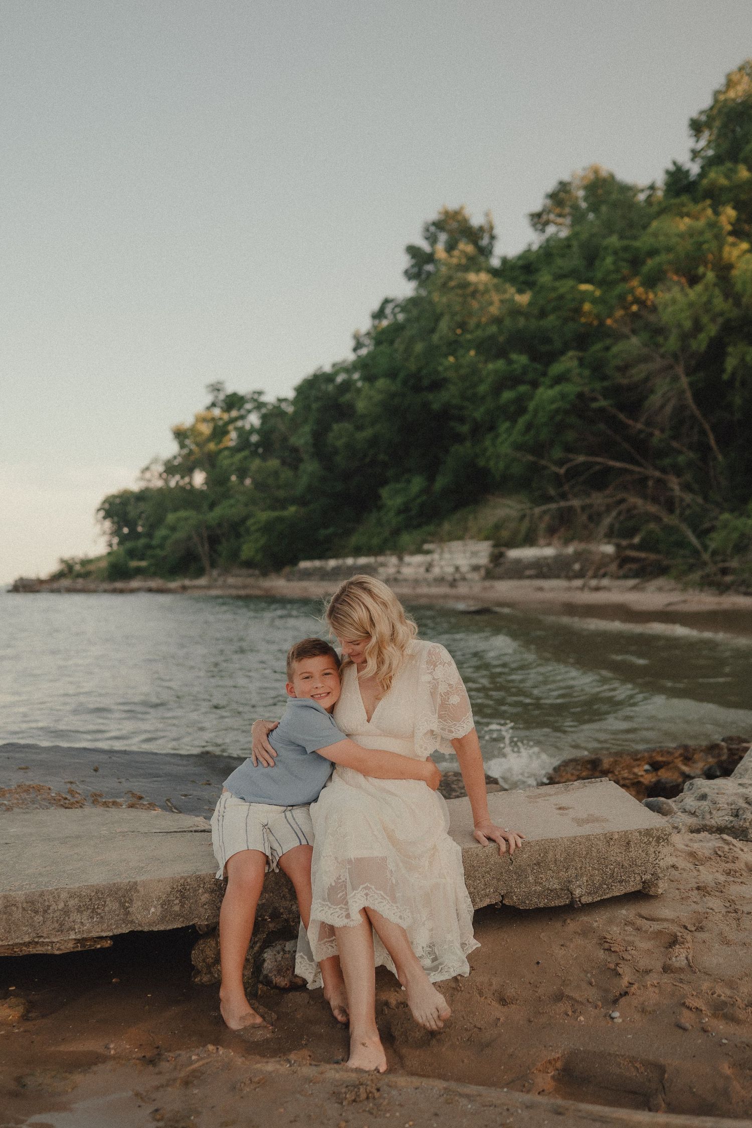 A smiling boy hugs his mother in a white dress as they sit together on rocks by a scenic beach.