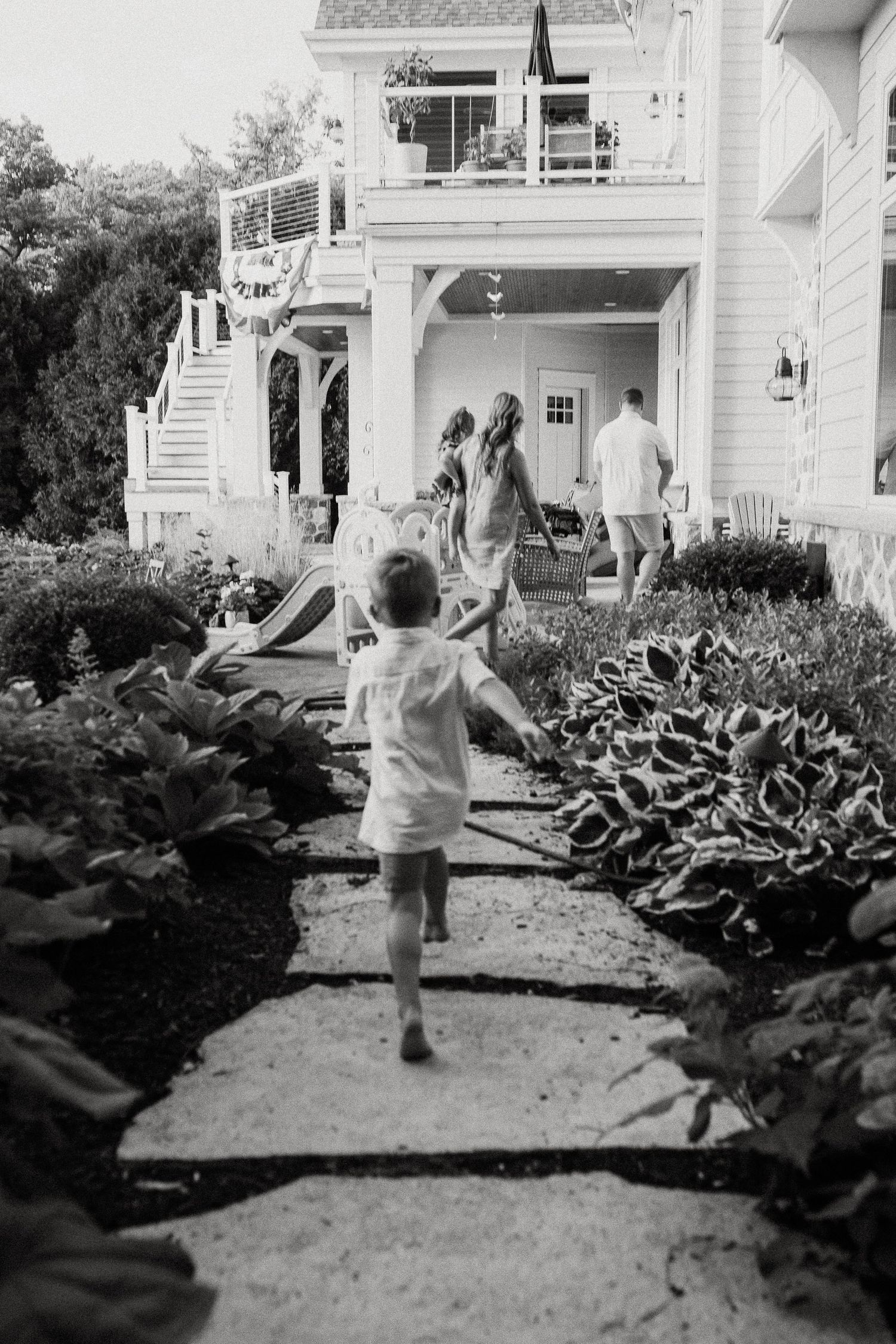 Black and white photo of a young boy running toward a white lakehouse along a garden pathway.