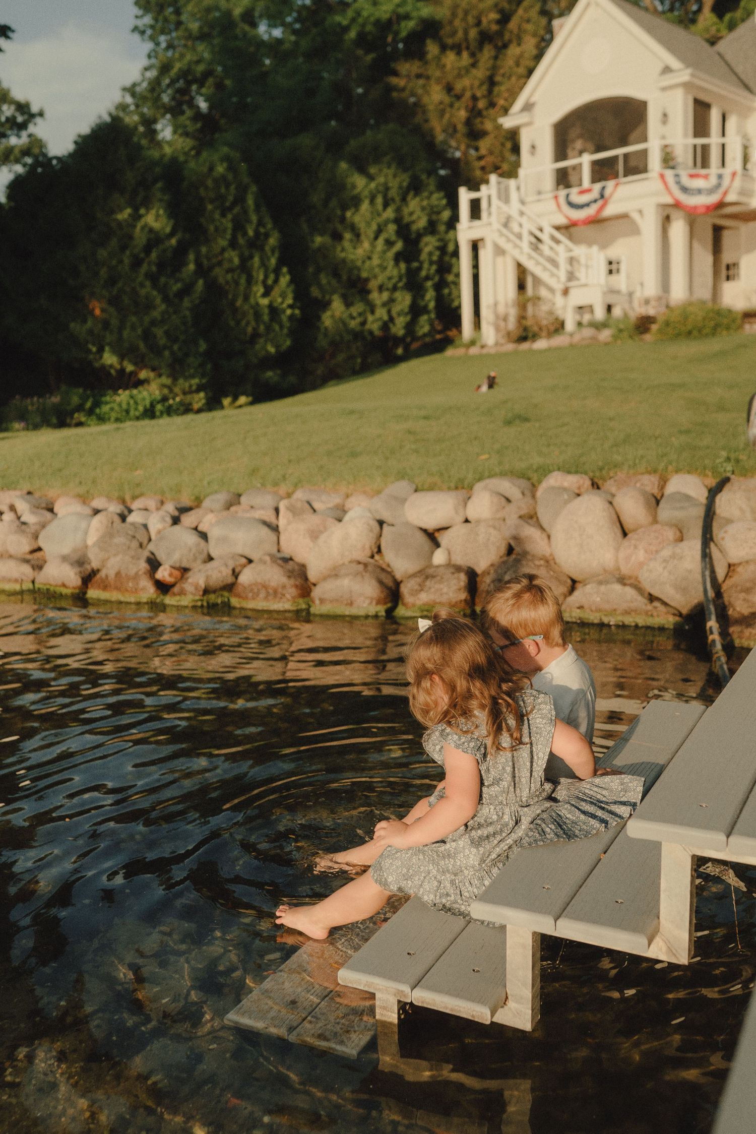 Two young children dangle feet off a dock into calm water near a lakeside home.
