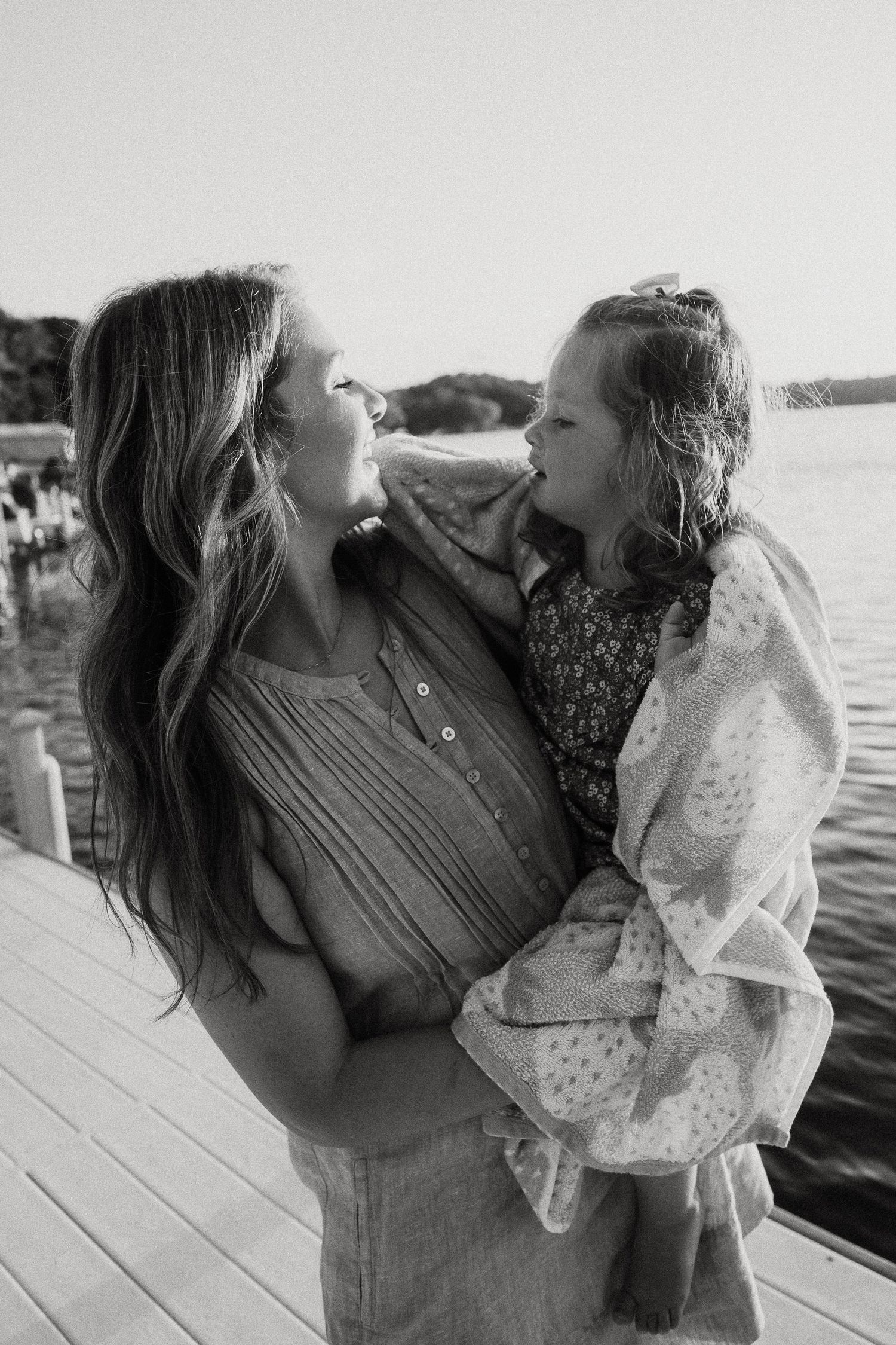 Black and white photo of mother holding young daughter sharing a sweet nose-to-nose moment on dock.