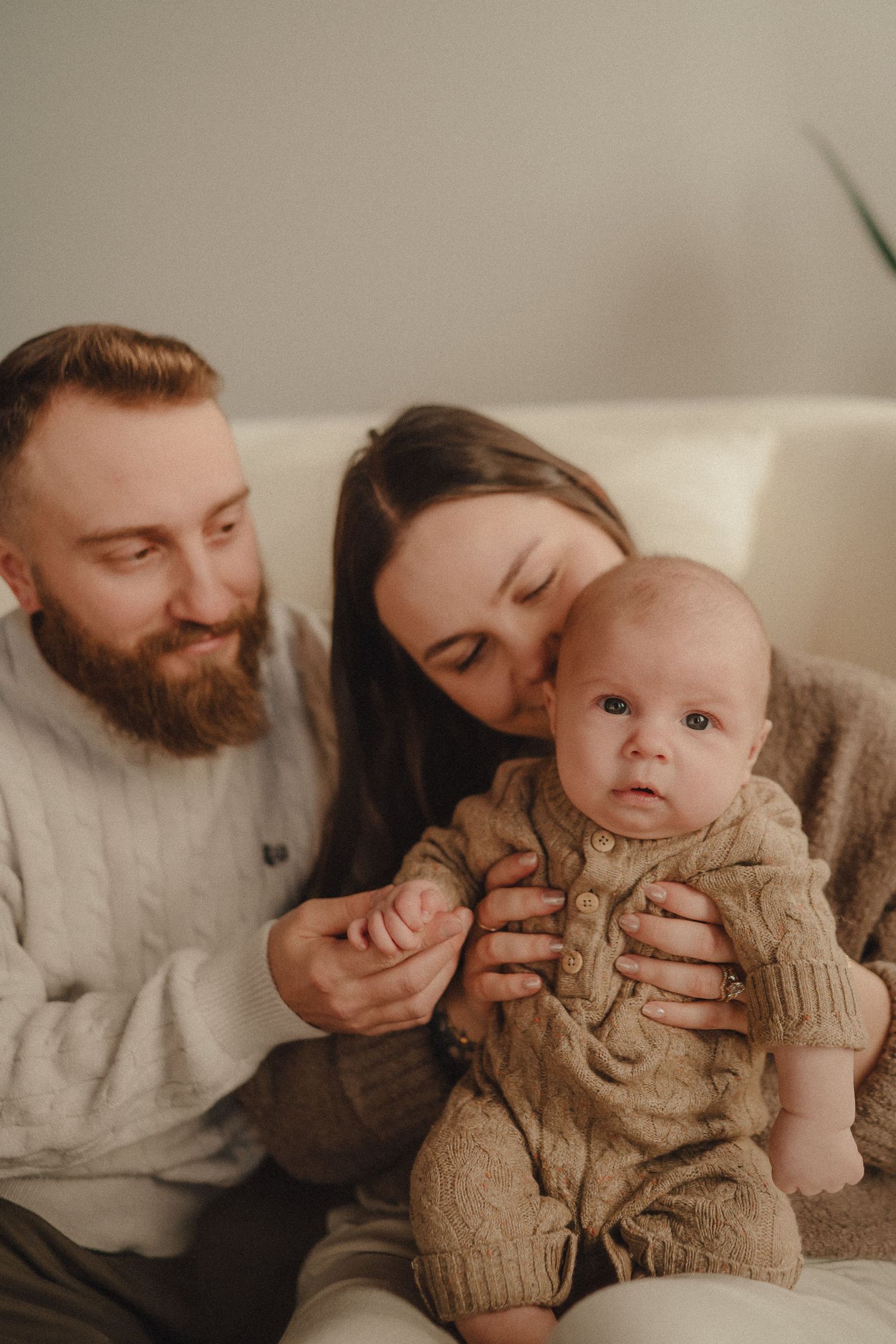 Indoor family photoshoot featuring a baby in beige knit clothing with parents in neutral tones.