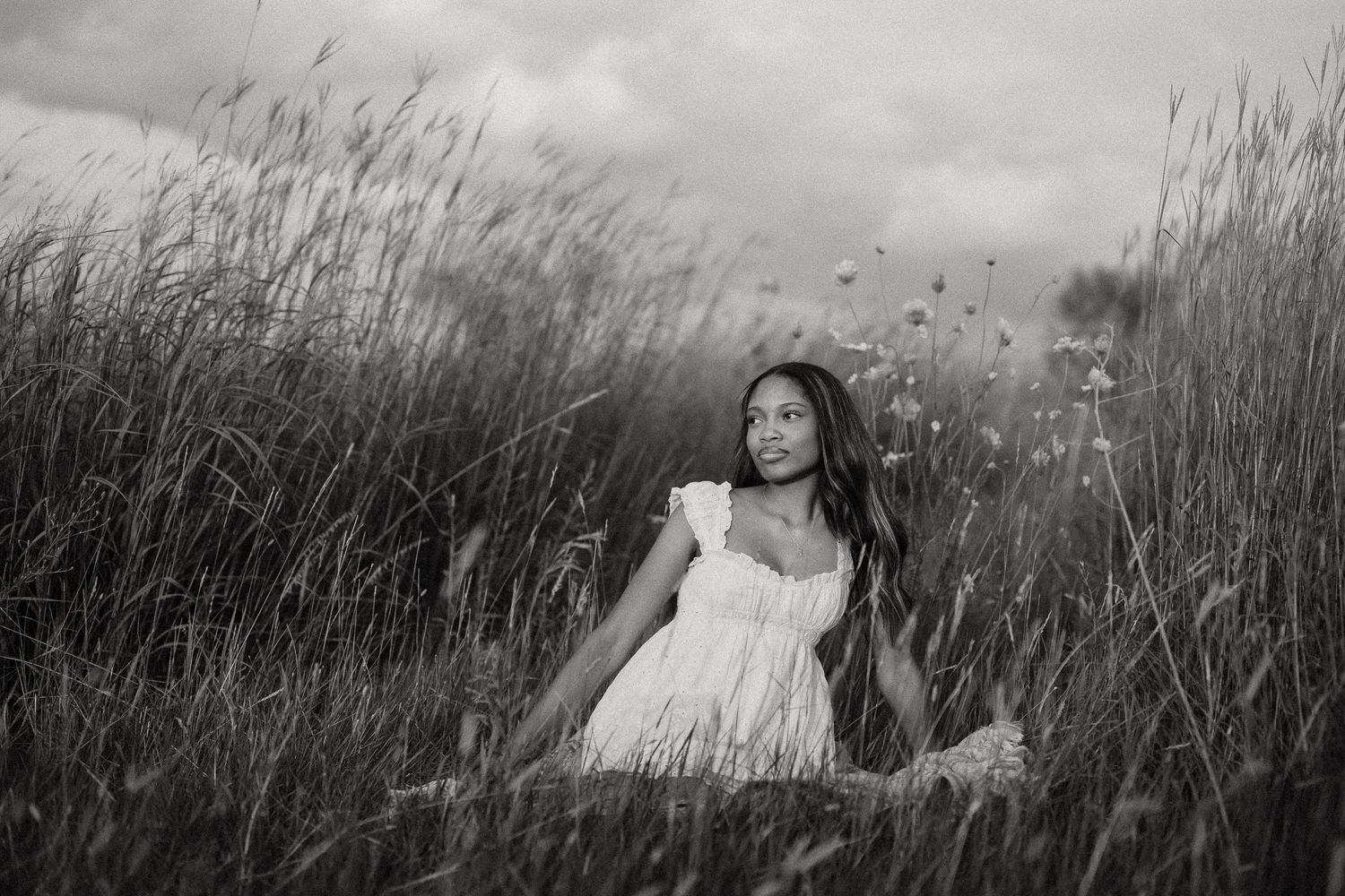 Black and white photo of a young woman in a white dress sitting peacefully among tall grass and wildflowers.