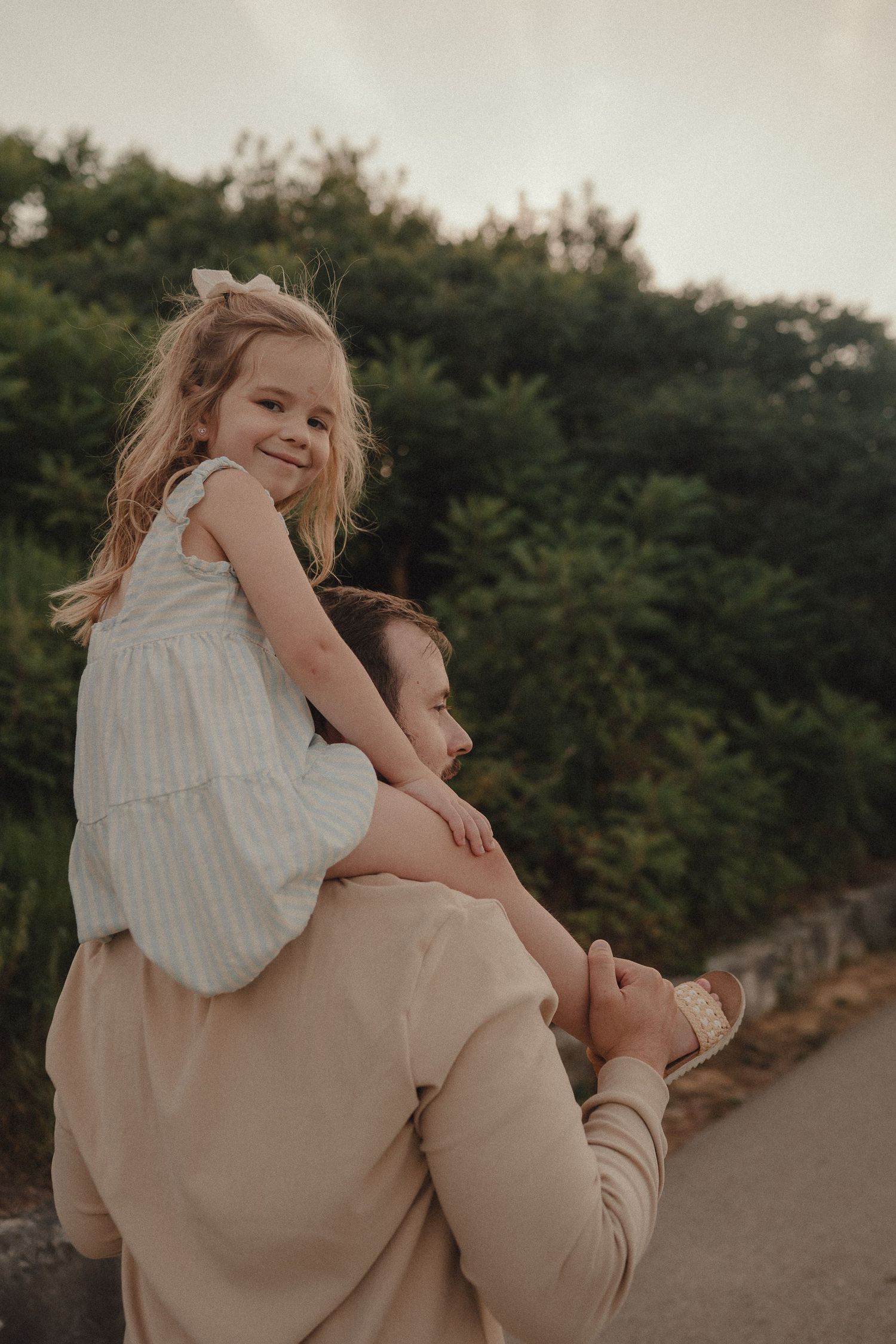 Young girl in striped dress sitting on fathers shoulders, smiling outdoors with green trees in background.