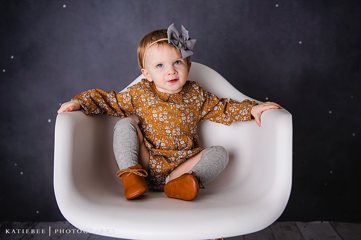 A toddler in a mustard floral dress and grey bow sits in a modern white chair against a dark backdrop.