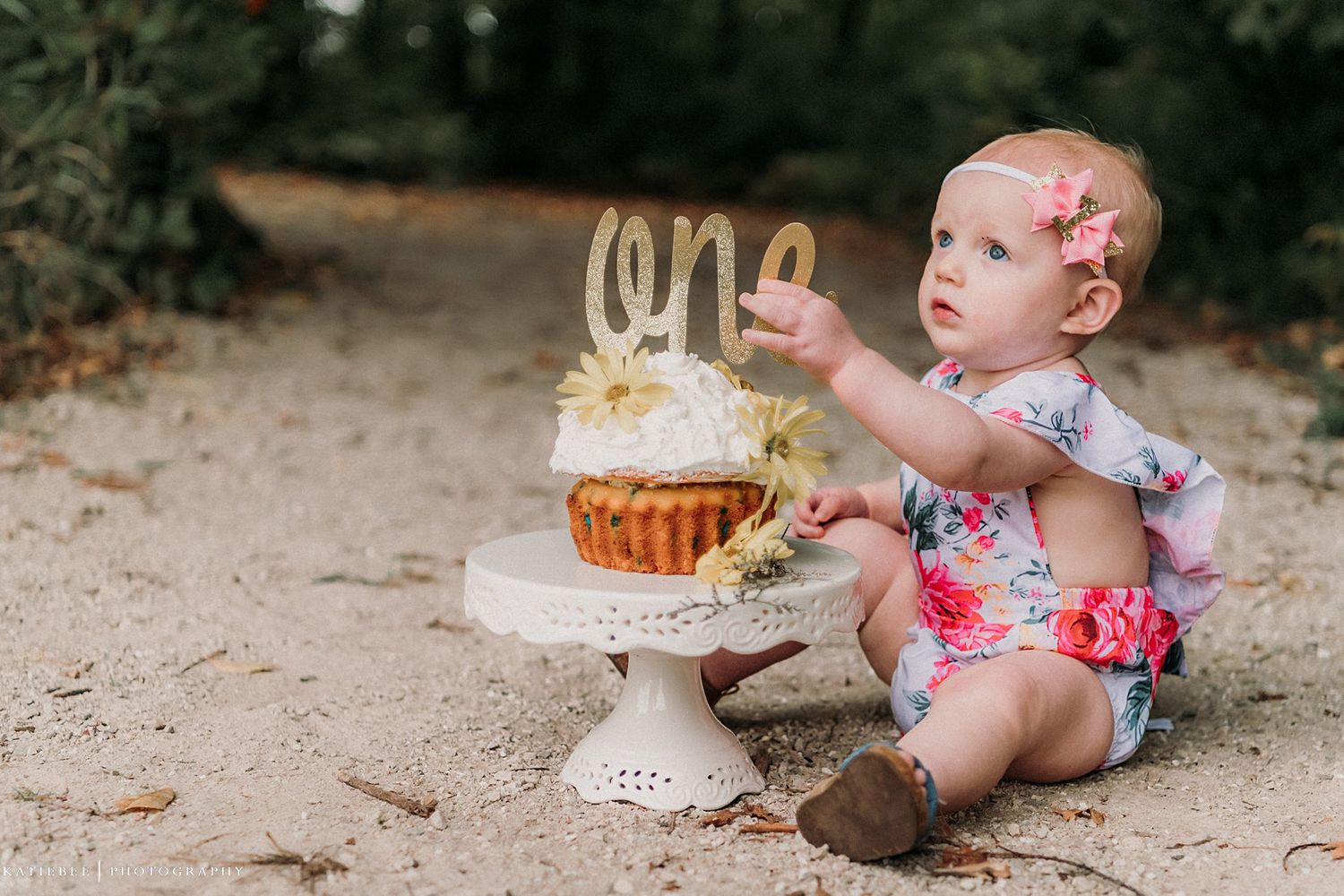 A baby in floral clothing reaches for a decorated cupcake on a pedestal.