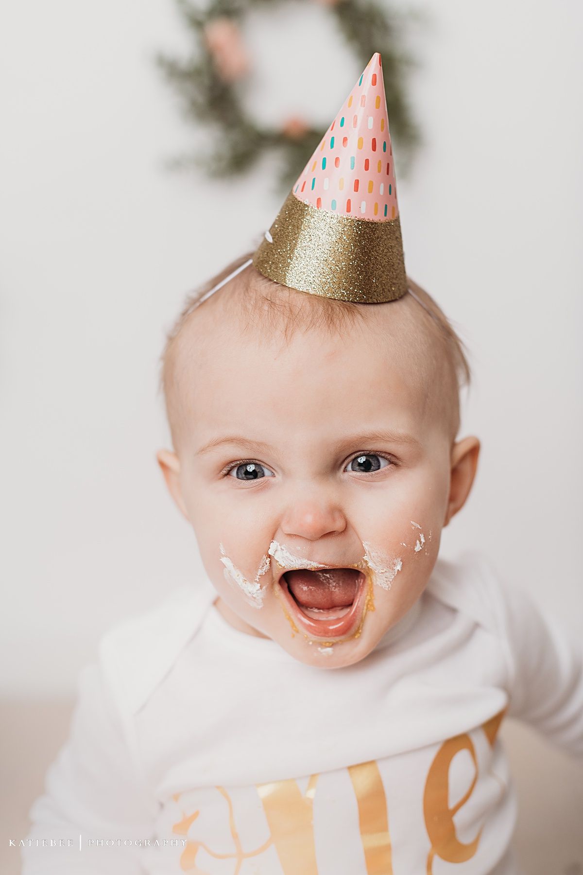 Baby wearing pink and gold party hat smiling with cake frosting on face.