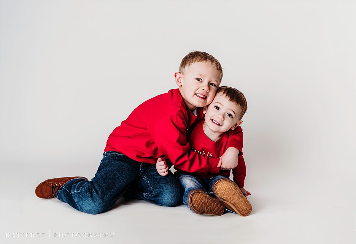 Two siblings in matching red sweaters hugging on white background.