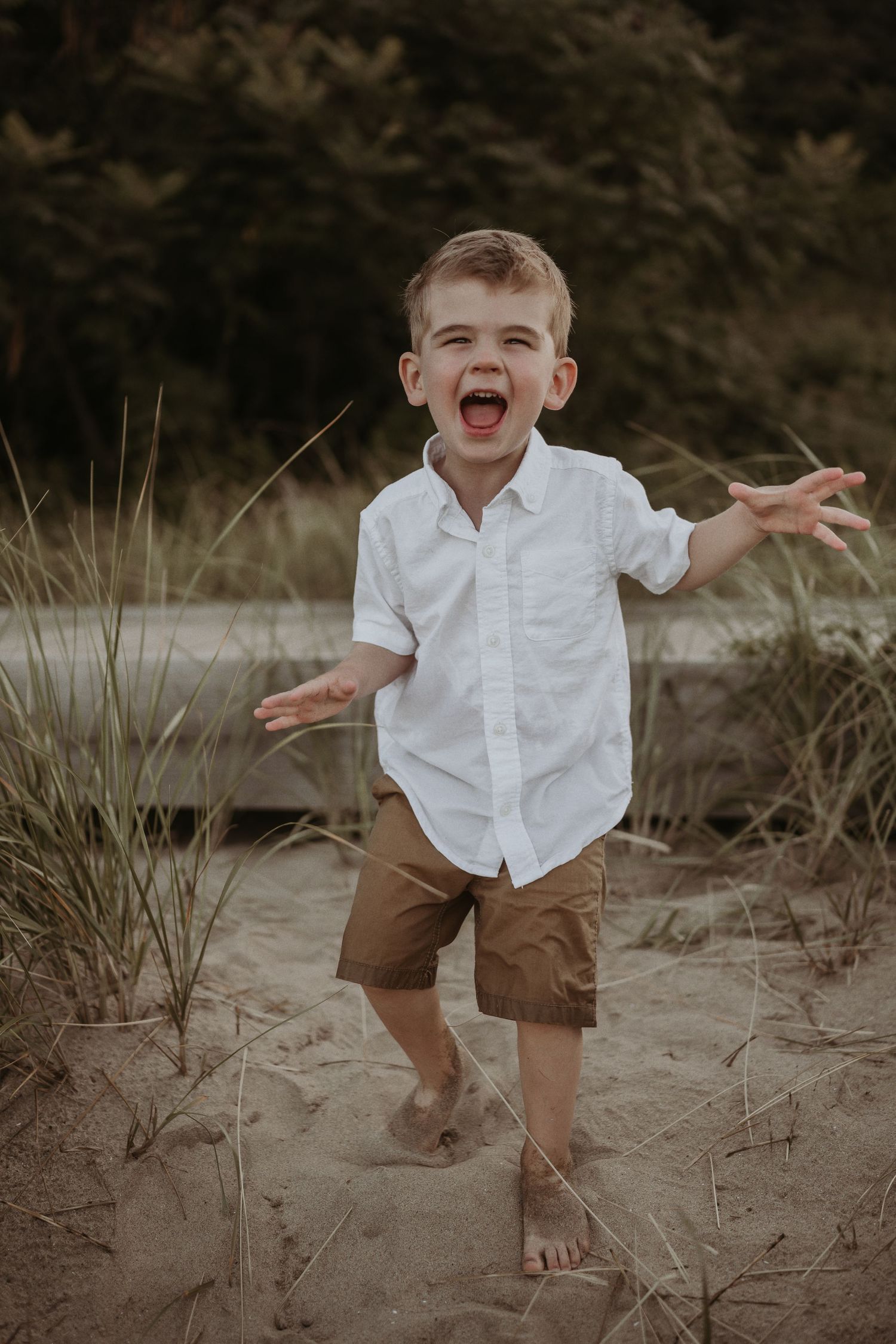 A joyful child in white shirt and brown shorts playing in beach grass.