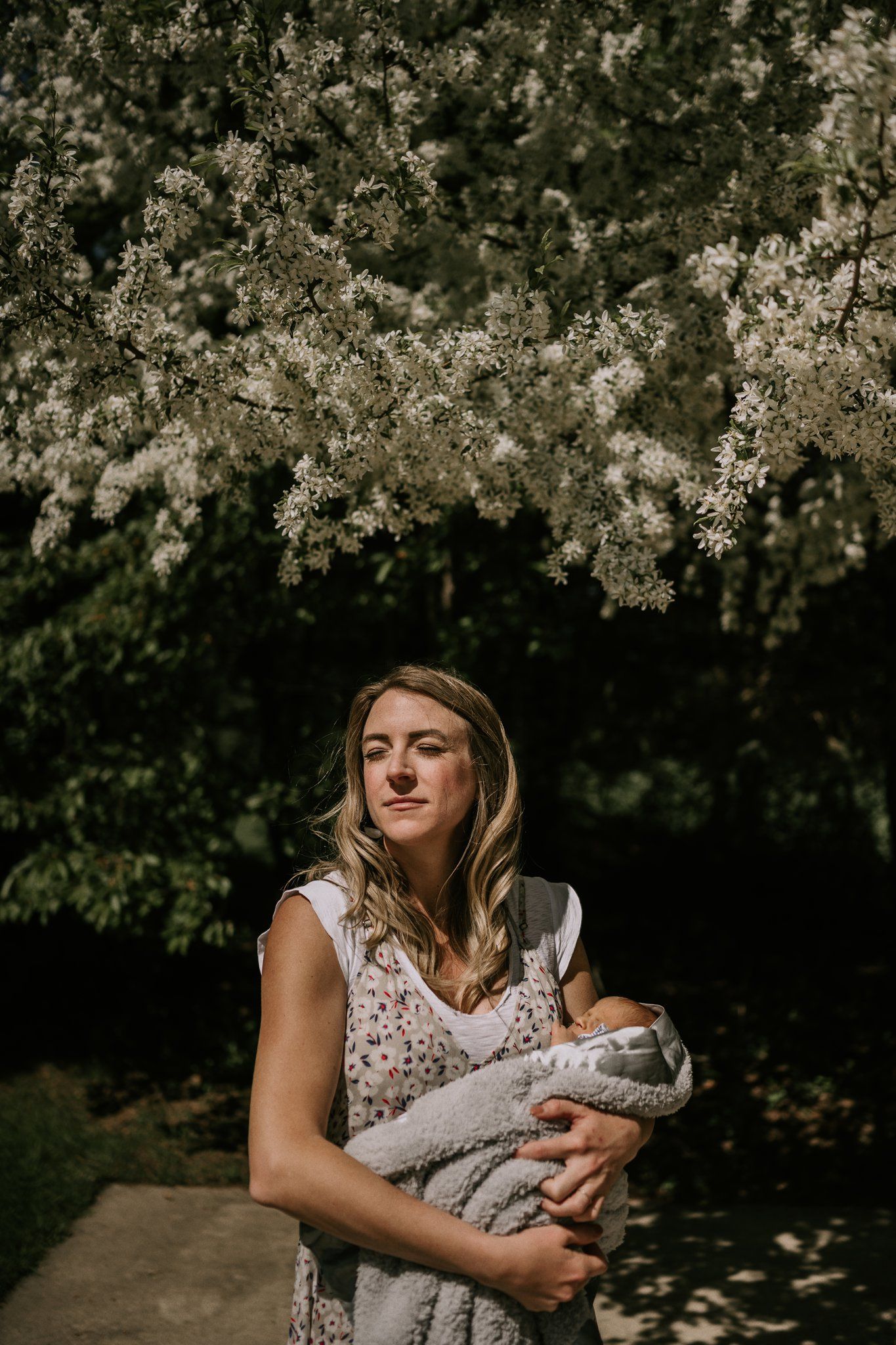 A person in a white lace dress holds a baby under blooming cherry blossom trees on a spring day.